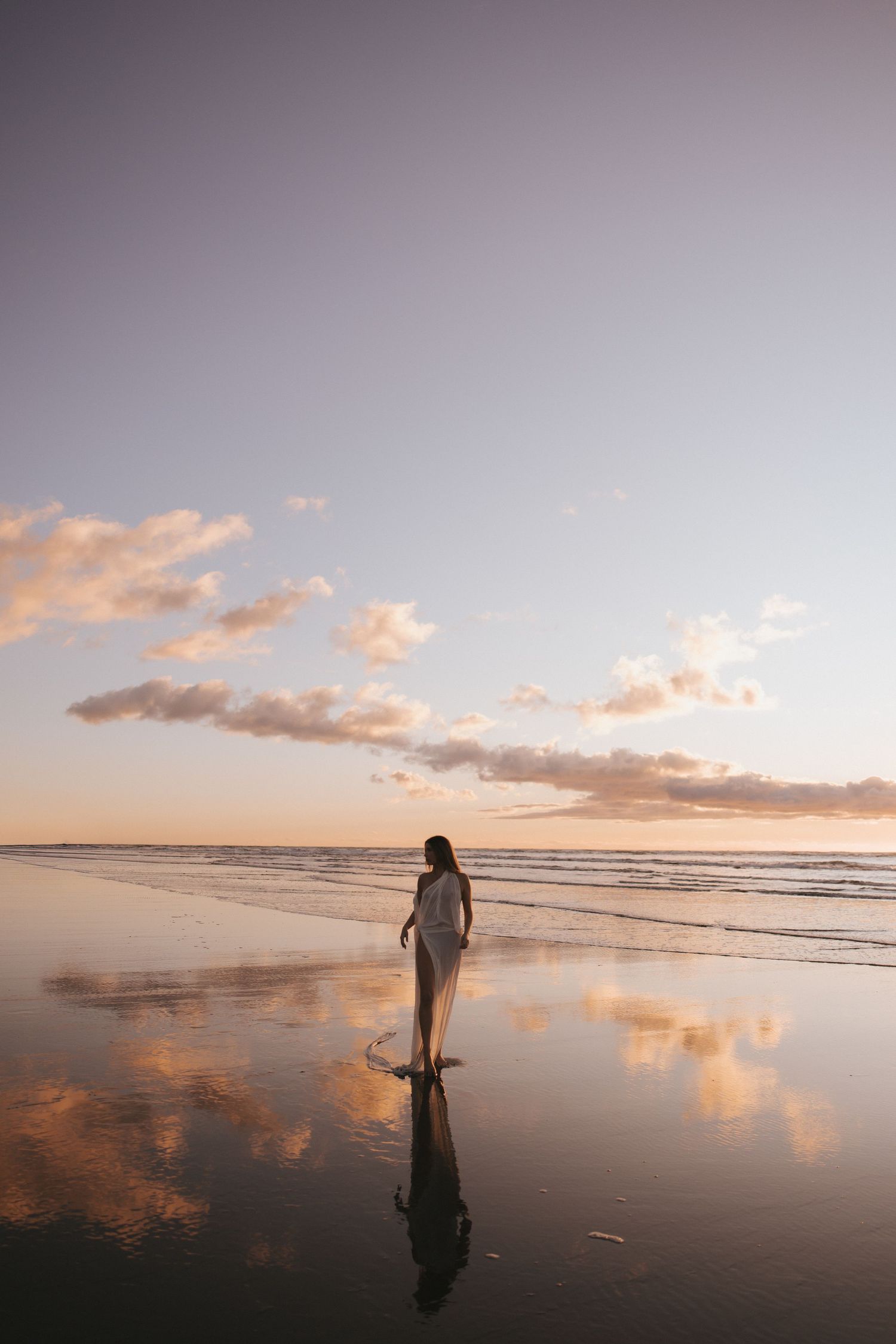 Woman in white dress walking on reflective beach at sunset in New Zealand.