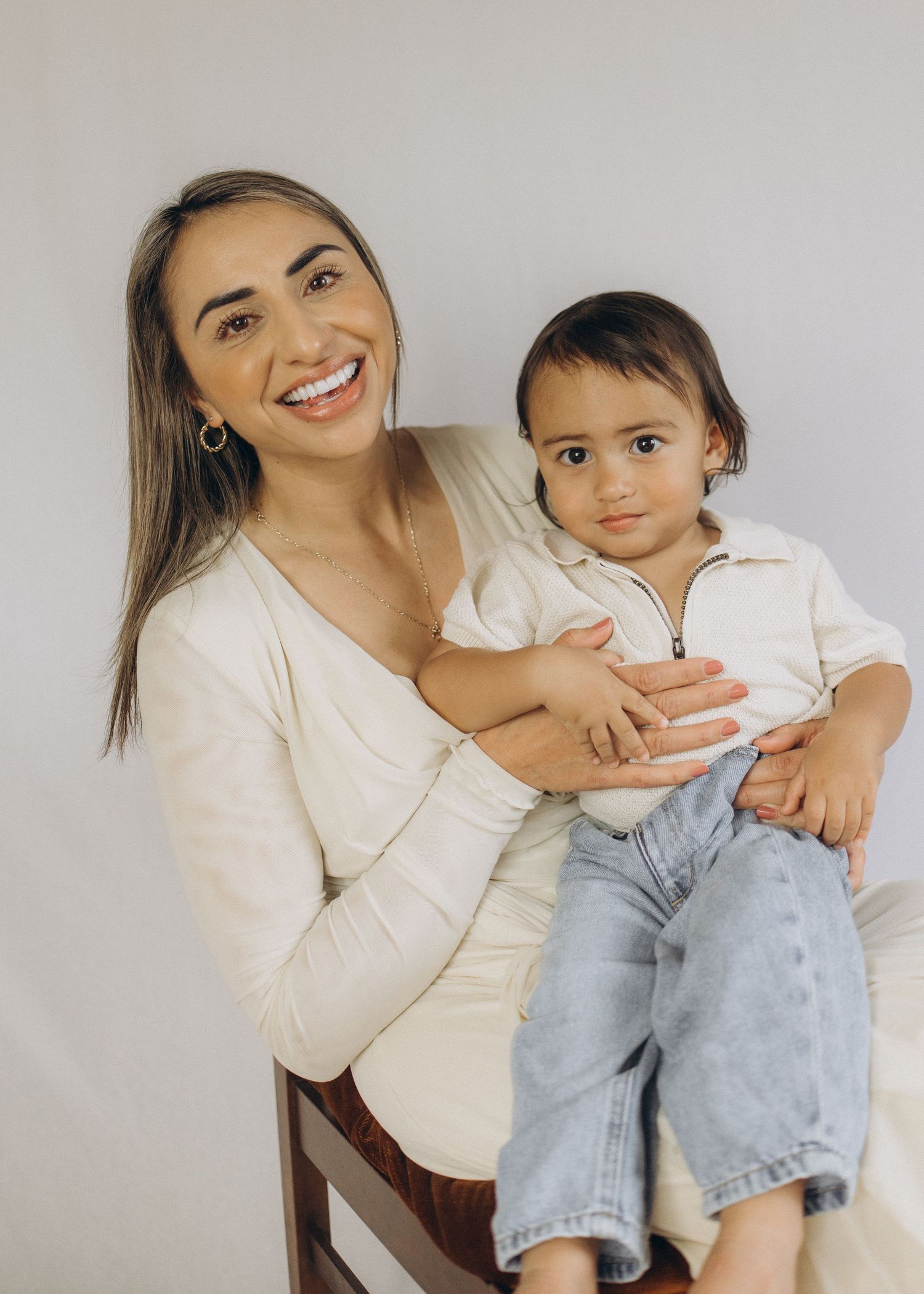 Smiling young woman in white top sitting on chair holding a toddler in jeans against a light background during a studio session in Raglan.