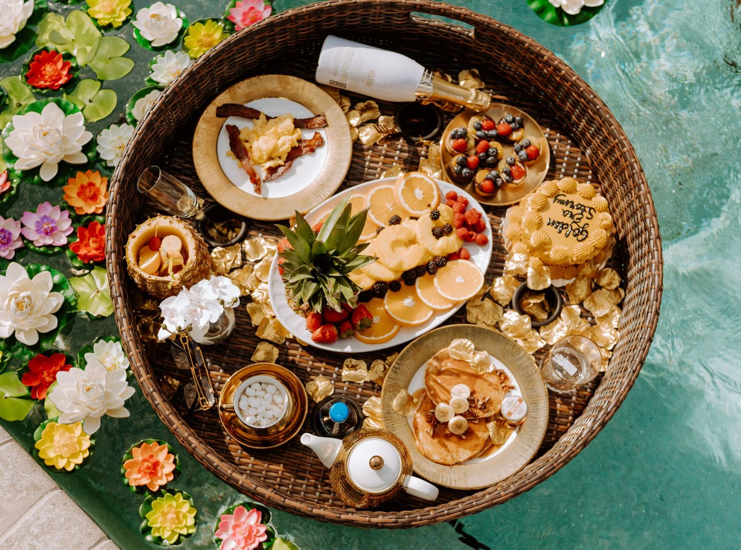 Overhead view of a wicker tray filled with pastries, desserts, and fresh berries on a turquoise background with flowers.