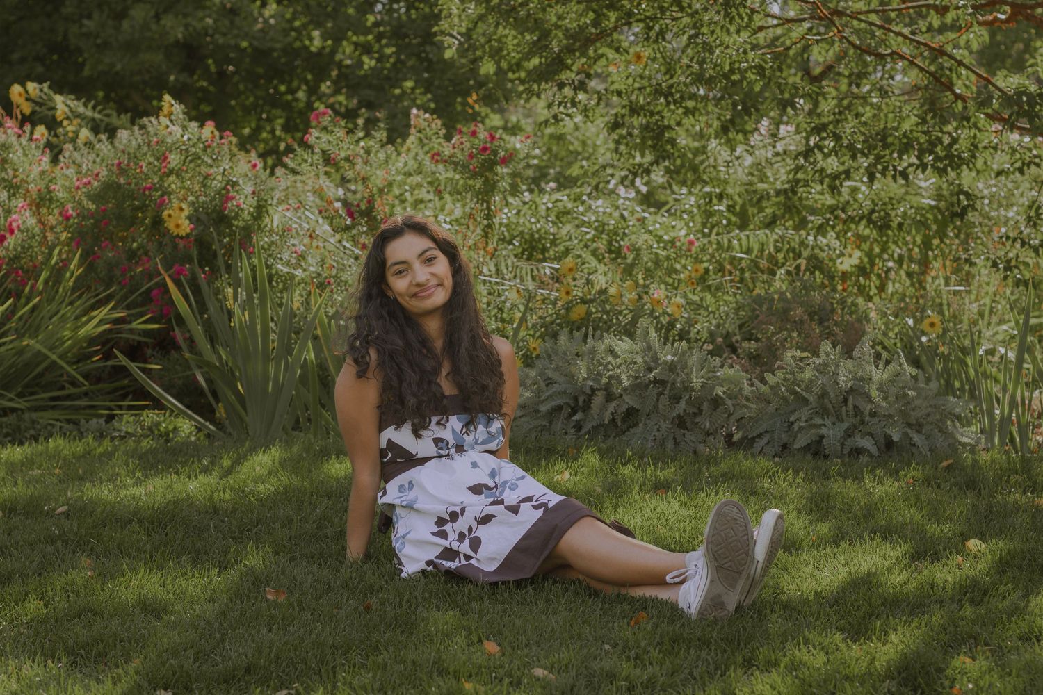 High senior girl sitting on lawn and smiling while wearing floral dress at Manito Park.