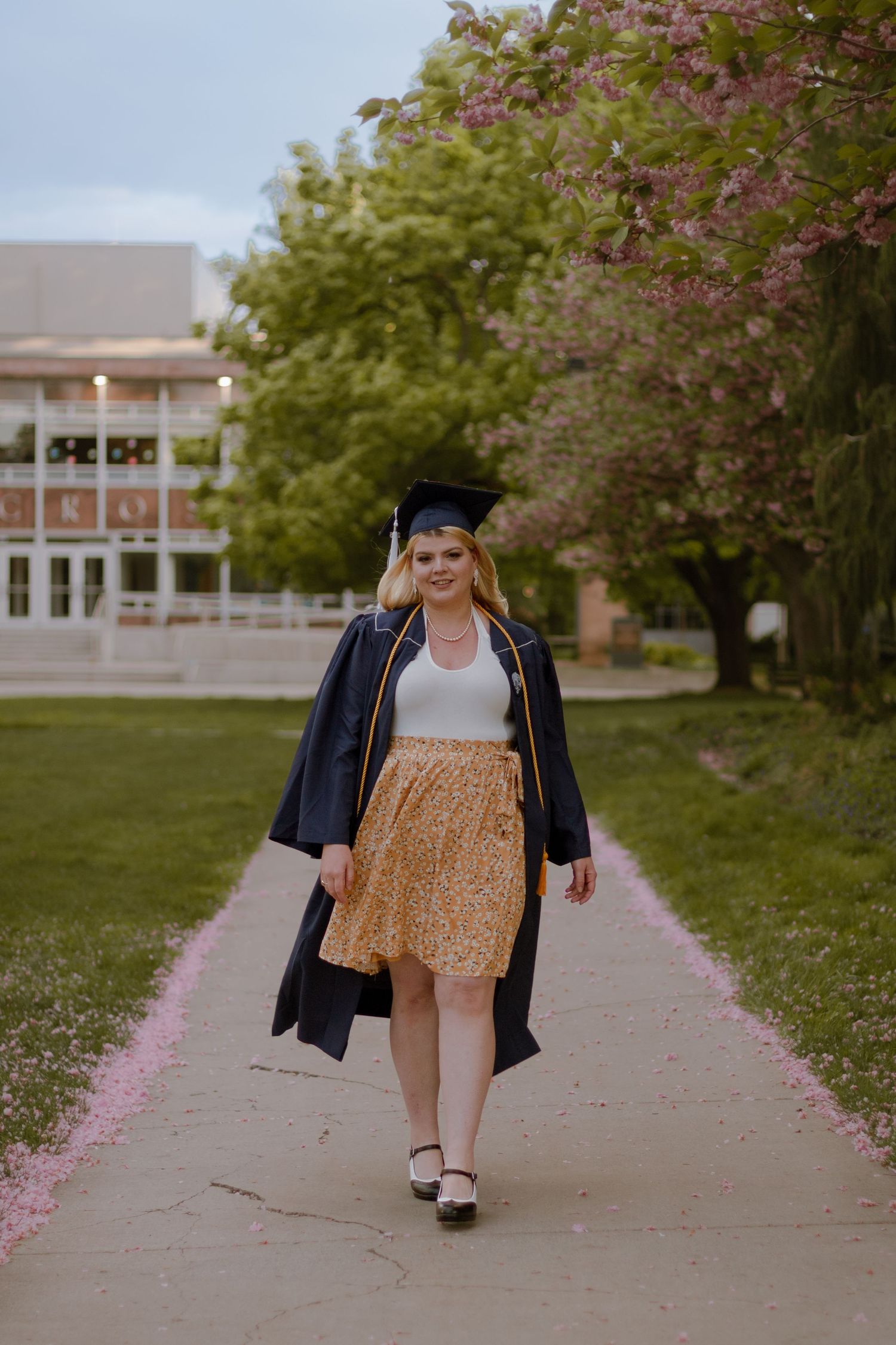 College graduate walking towards photographer on campus near cherry blossom trees.