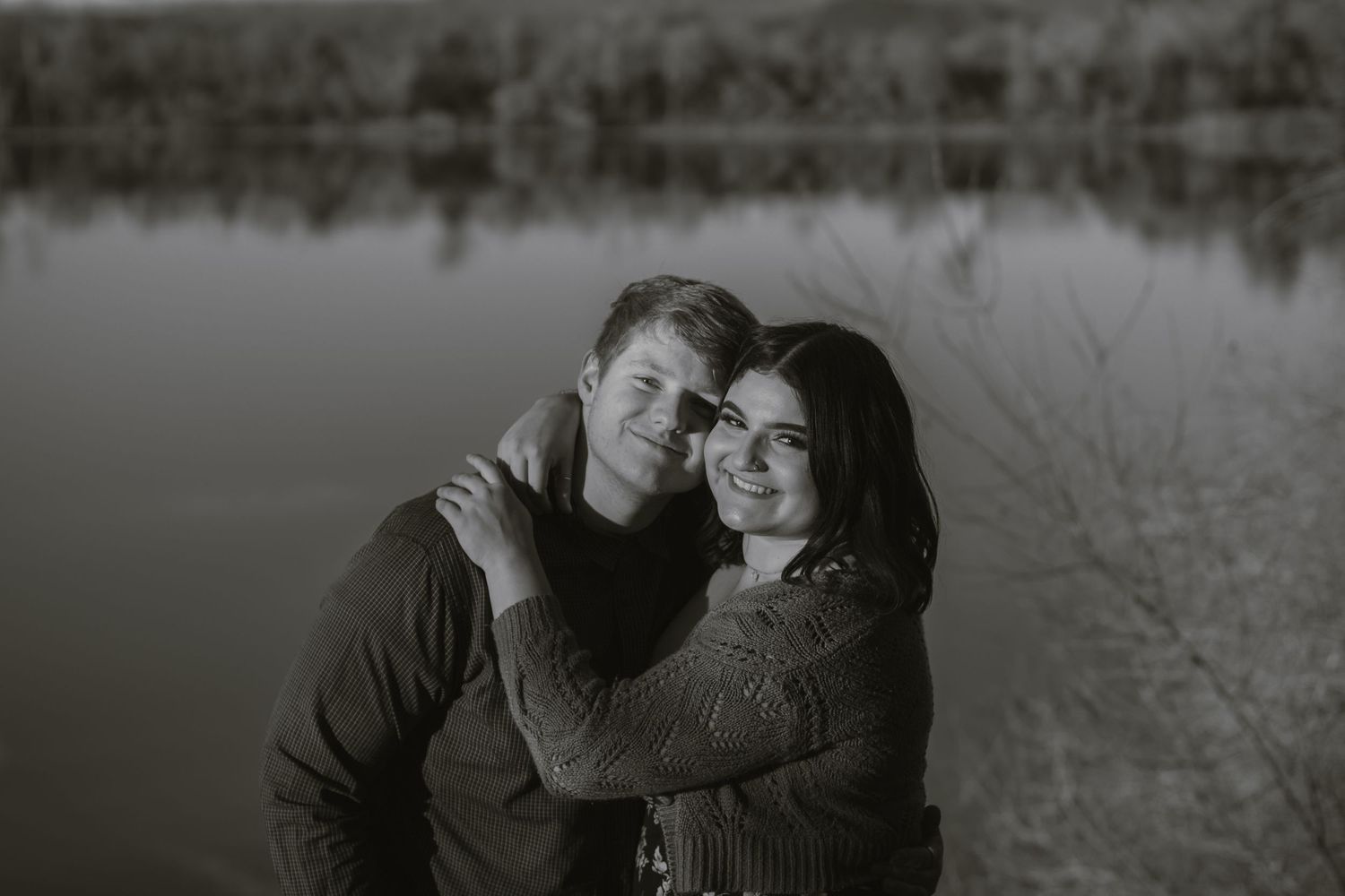 Woman and a man romantically involved posed hugging in front of lake reflecting the trees and brush surrounding it at Riverfront Park, CA.