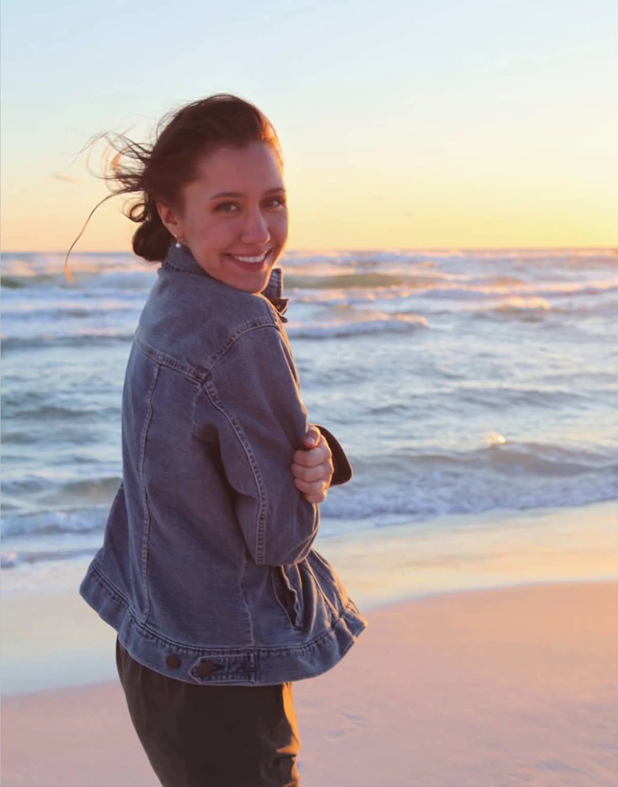 Smiling woman in a denim jacket looks back over her shoulder on a windswept beach as the sunset glows over the ocean waves.