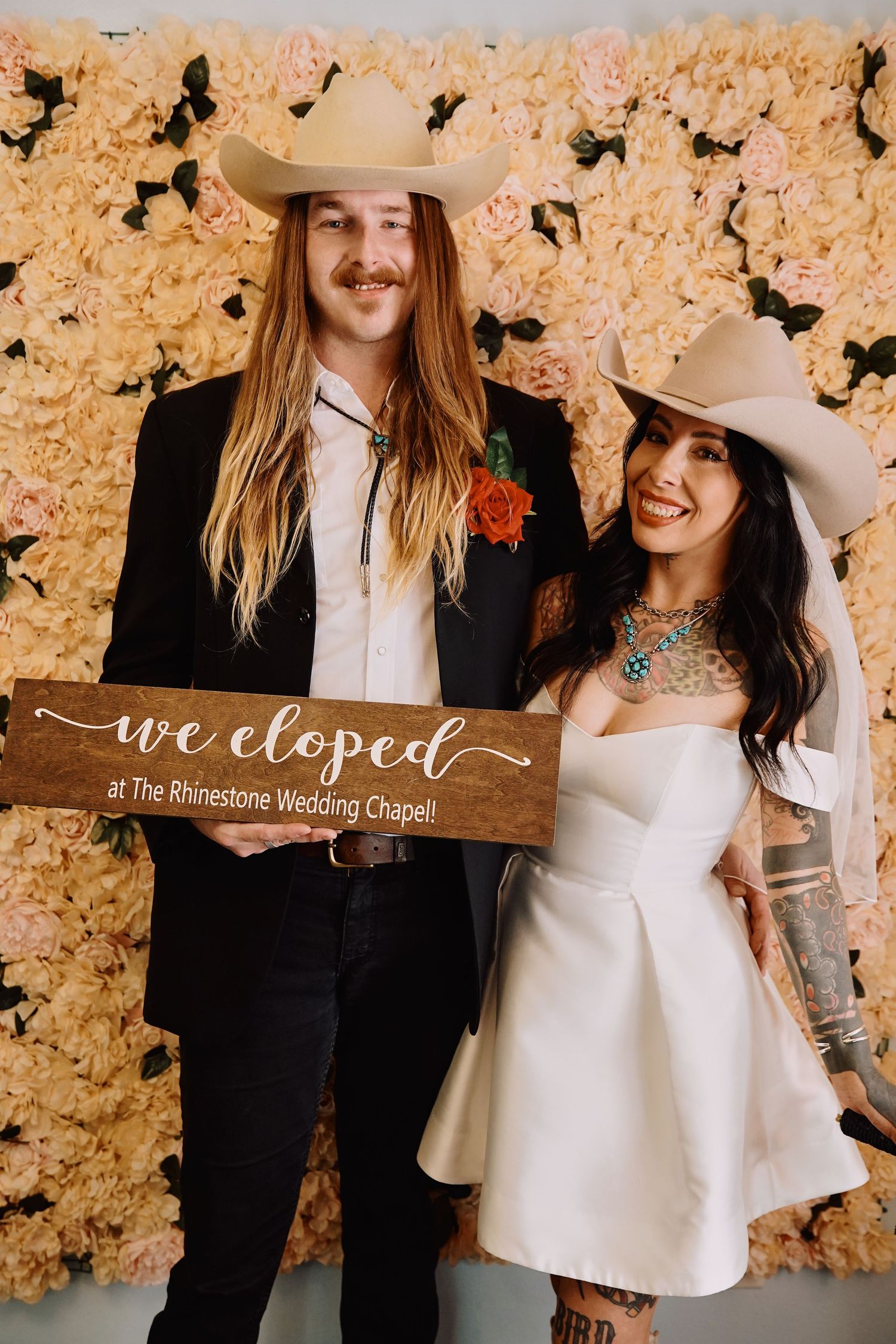 A couple poses together in front of a floral wall with a rustic wooden sign at The Bellissima Wedding Chapel.