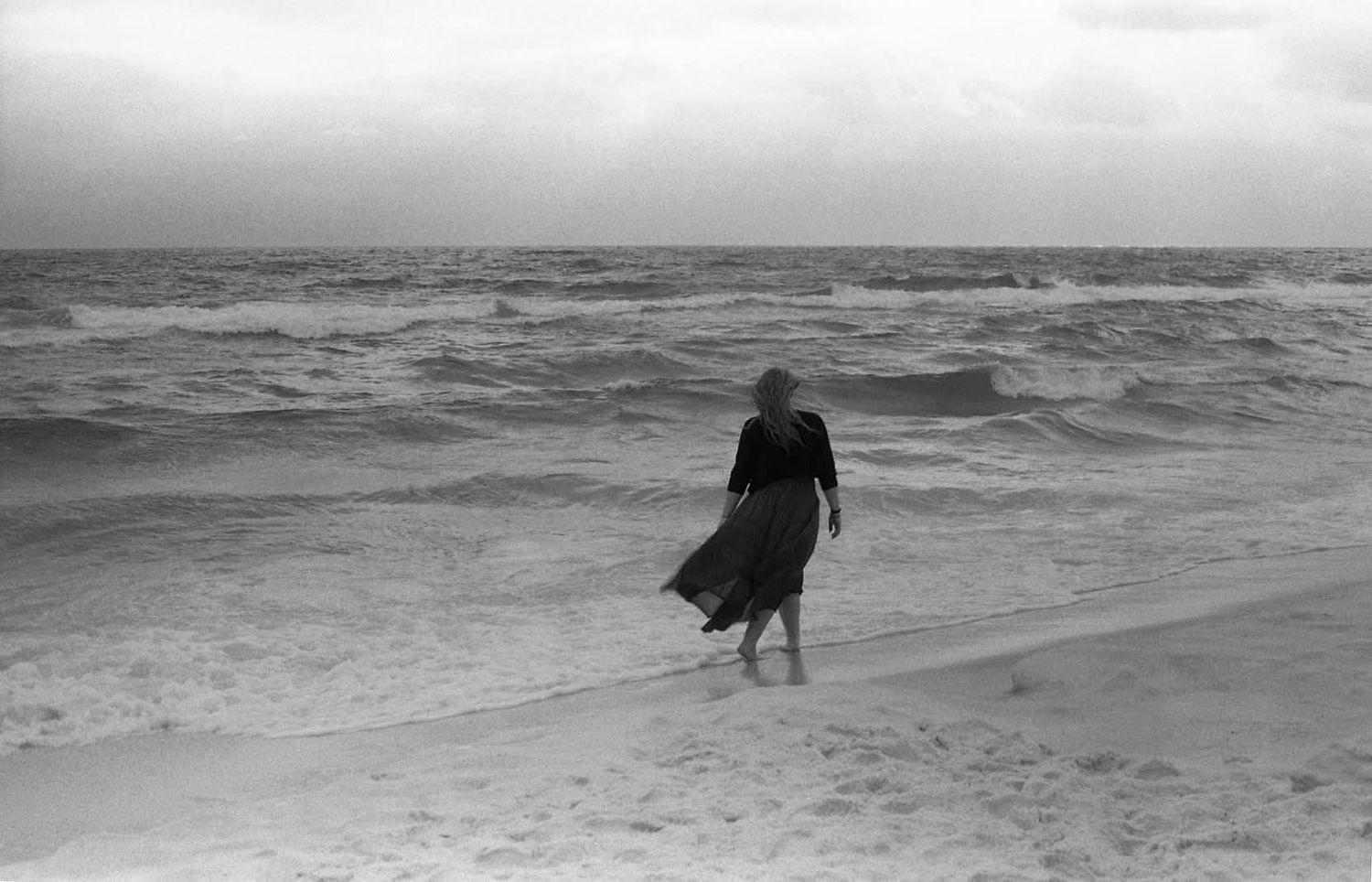 Black and white photo of a woman in a flowing dark dress walking barefoot along the shore as ocean waves roll in under a cloudy sky.