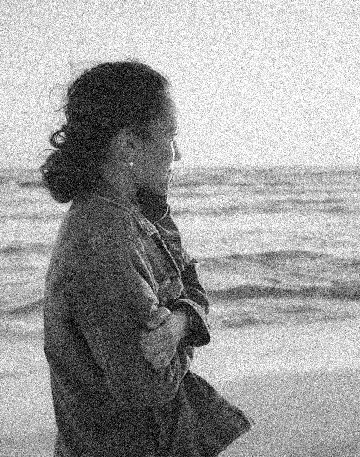 Black and white photo of a woman in a denim jacket standing on the beach, arms crossed, gazing at the ocean waves.