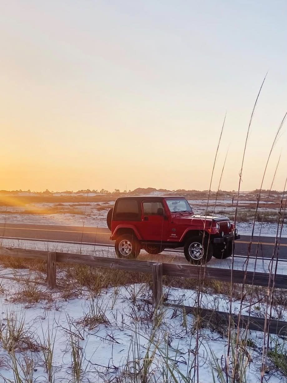 Red Jeep Wrangler parked on a road beside a wooden fence, with snow-dusted dunes and tall grasses glowing in golden sunset light.