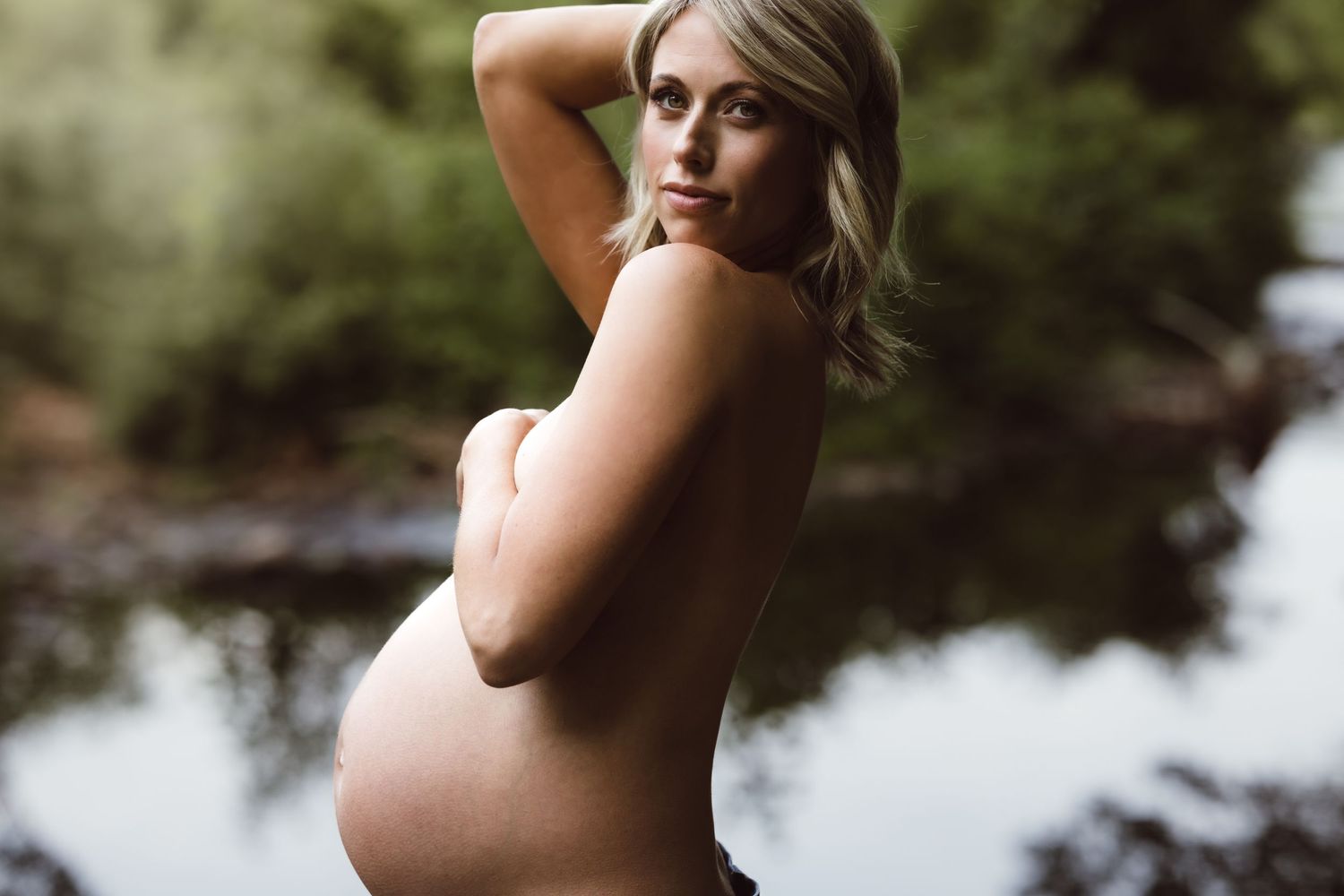 A silhouetted pregnant figure poses in profile against a tranquil lake backdrop during a maternity photoshoot.