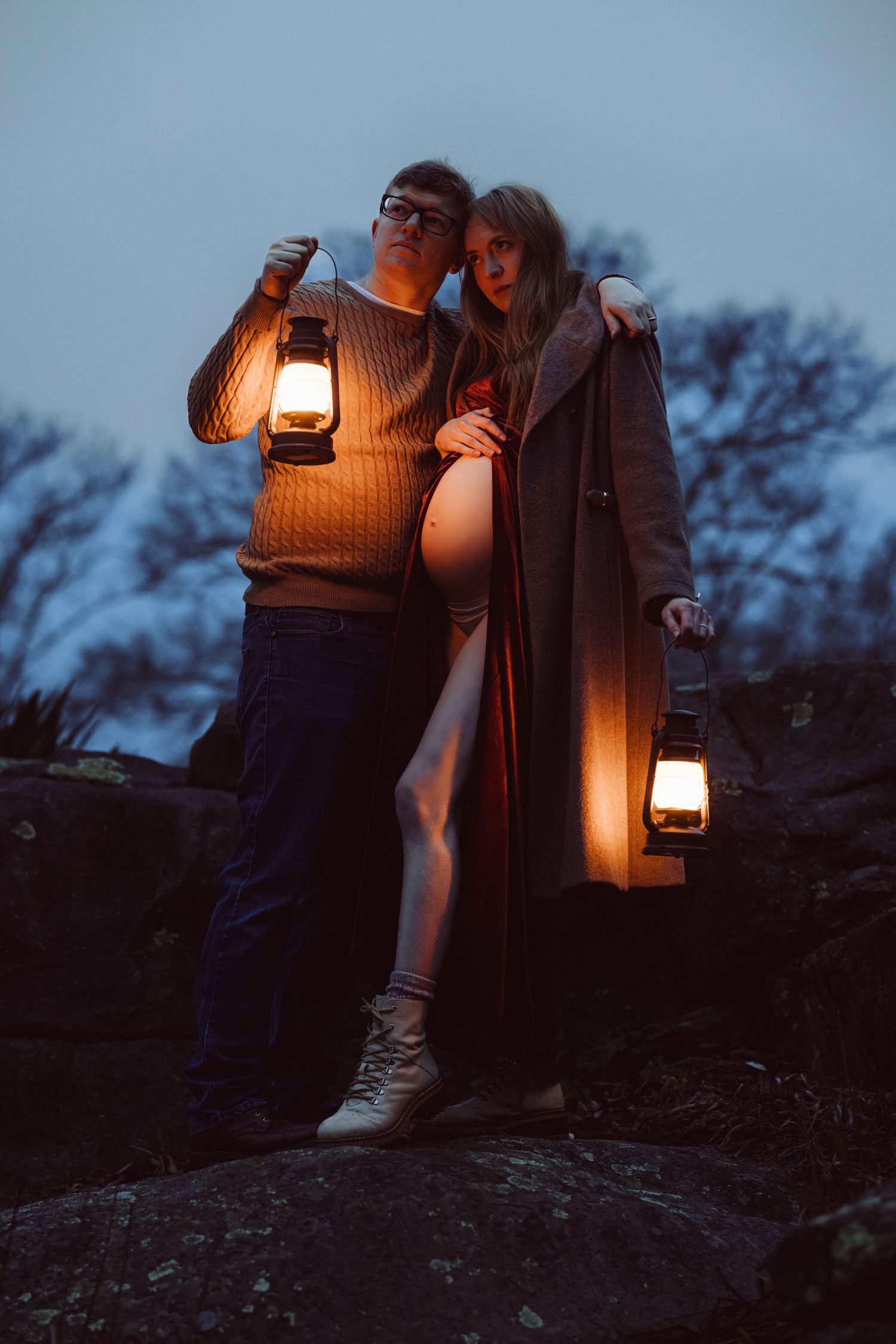 A pregnant couple holding lantern illuminates a dark outdoor scene at dusk on a giant rock for maternity photoshoot.