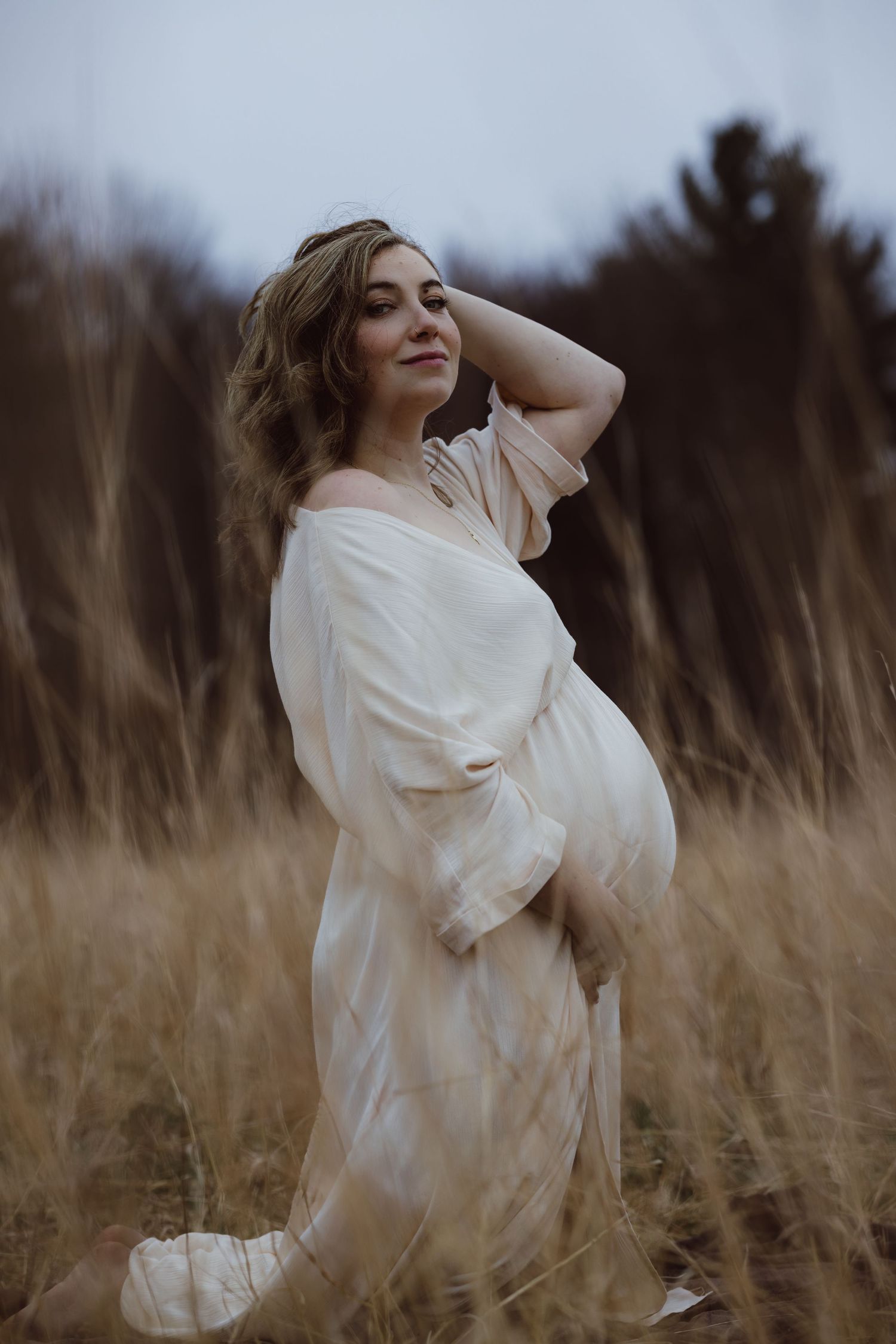 A pregnant mother in a flowing white dress poses gracefully in a Connecticut field of tall dried grass at dusk for maternity photoshoot.