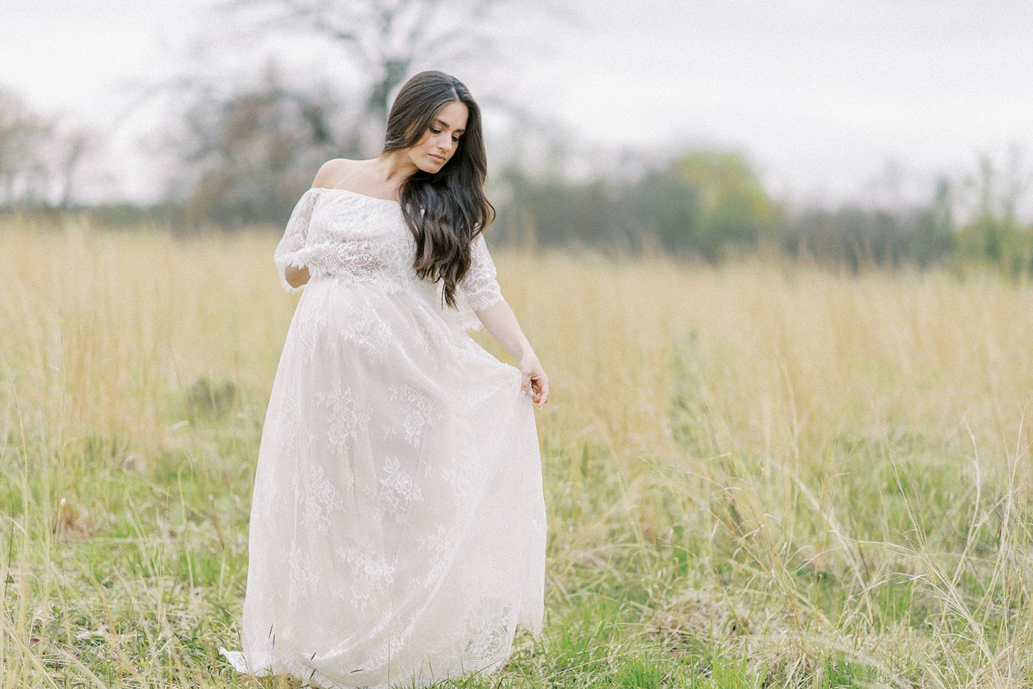 A mother in a flowing white dress walks through a sunlit meadow of tall grass and wildflowers.