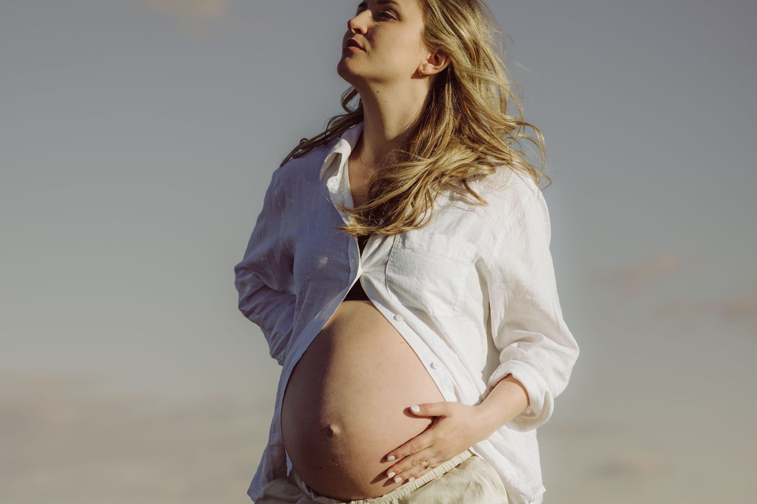 A pregnant woman in white clothing holds their bare belly while the wind blows through her hair against a sunset sky at a Westport beach.