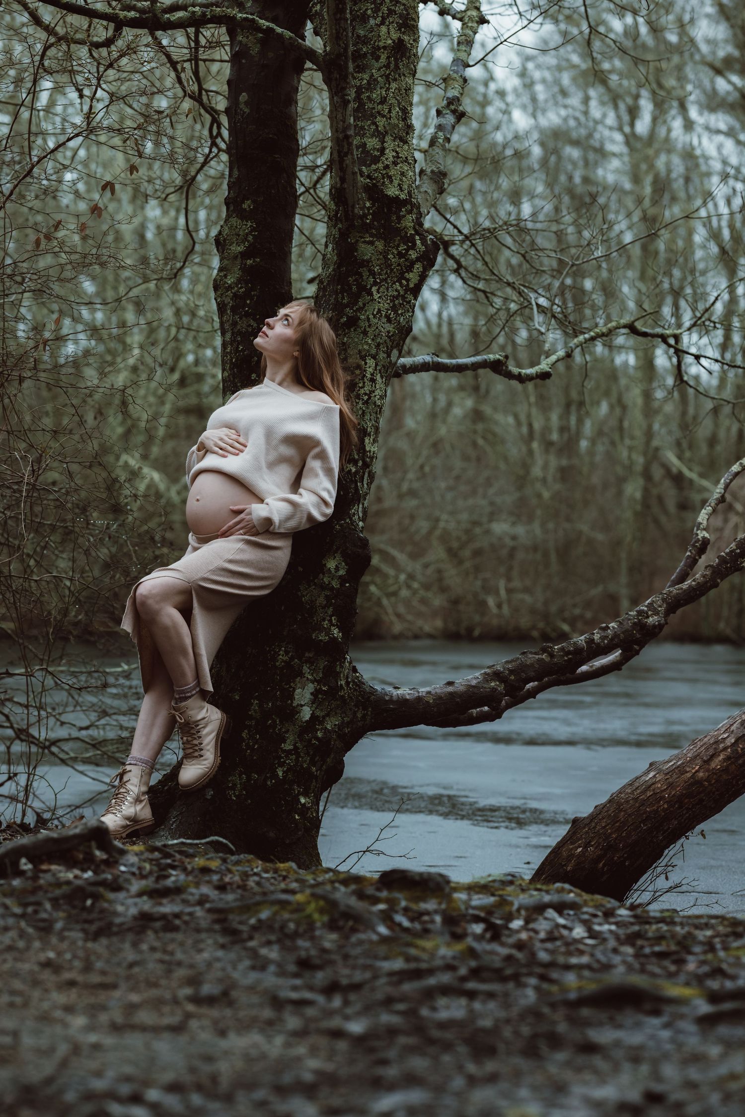 A pregnant woman leaning on a tree in the forest by a frozen pond posing for a maternity session in winter.