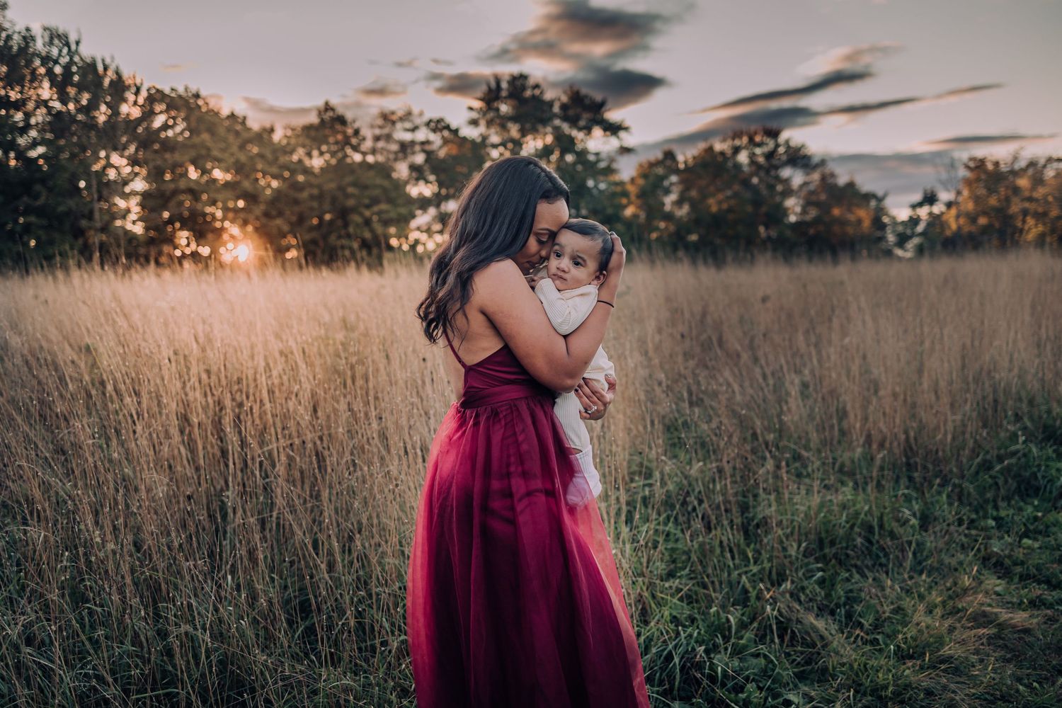 A mother in a flowing burgundy dress stands in a golden field at sunset while holding a small child.