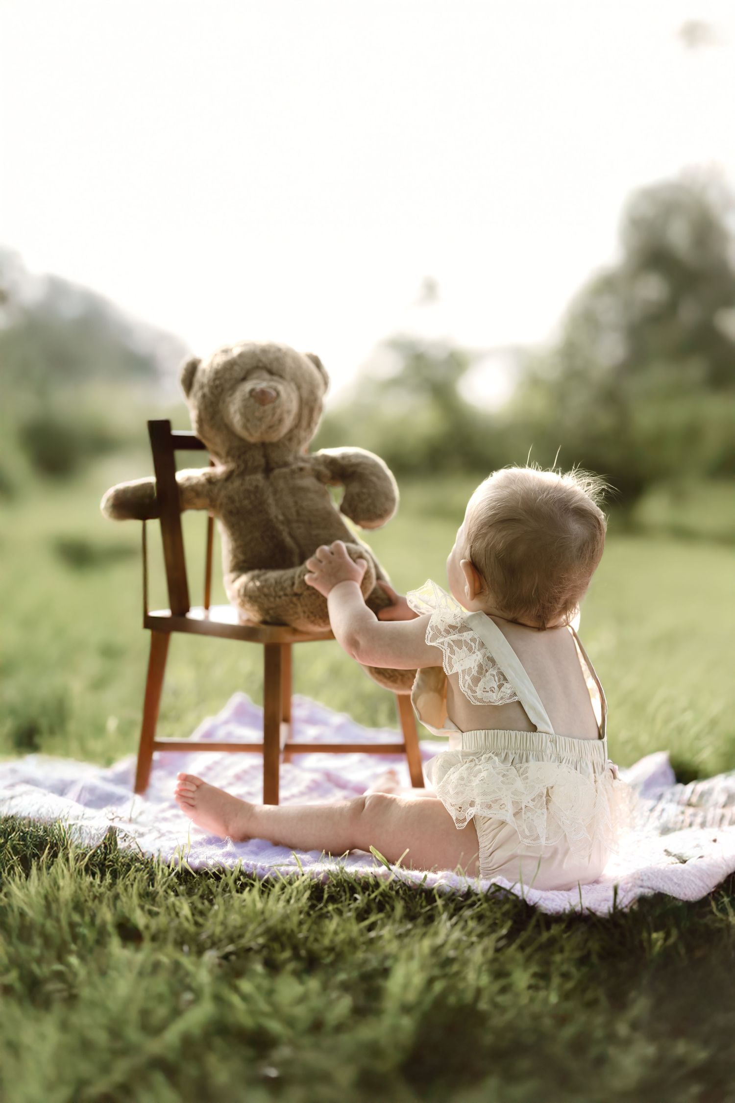 A one year old girl in lace onesie reaching for her favorite teddy sitting on a chair outdoors at sunset for milestone photoshoot.