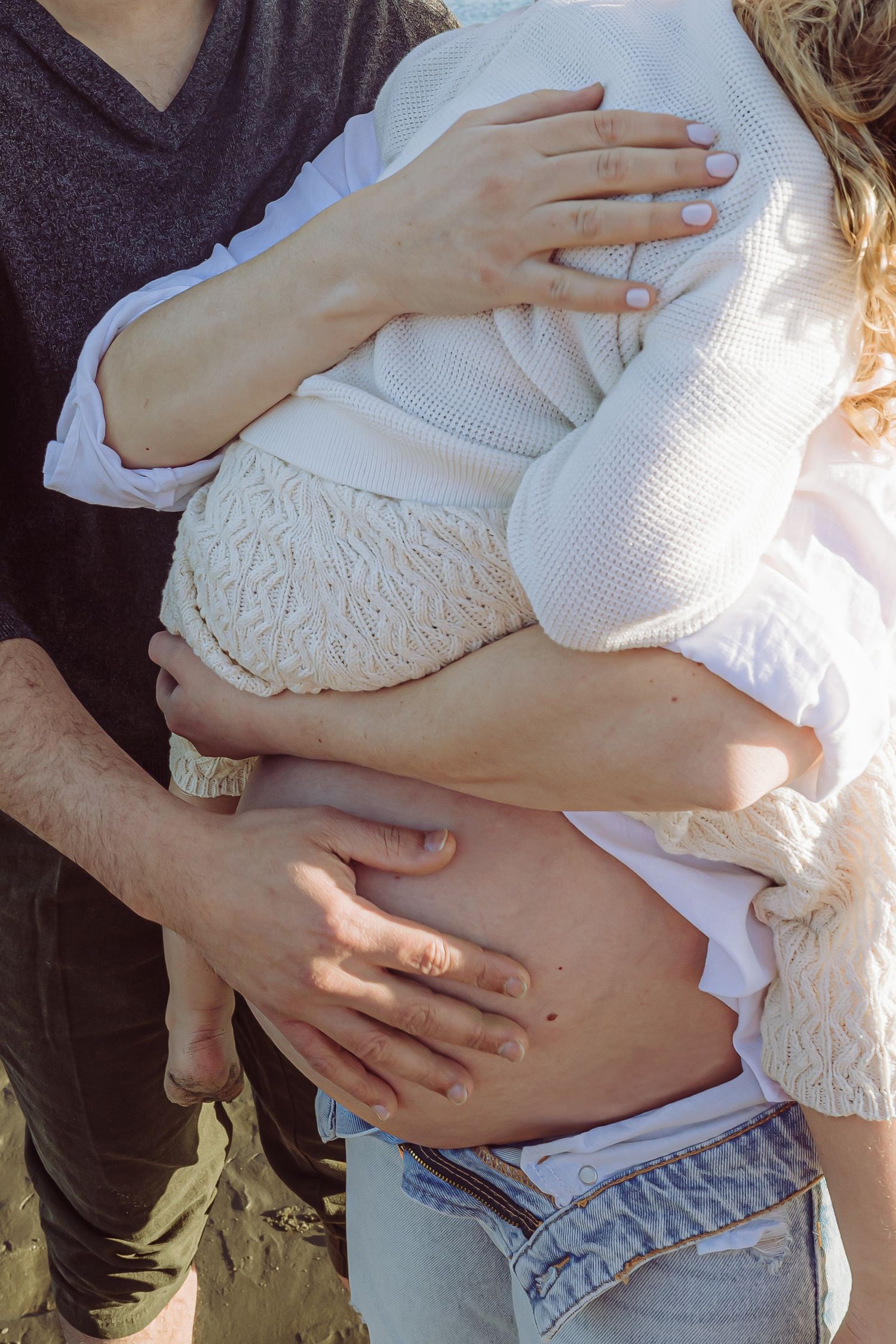 A couple embraces while showing a pregnant woman's bare belly in casual clothing with sunlight glowing in the background.