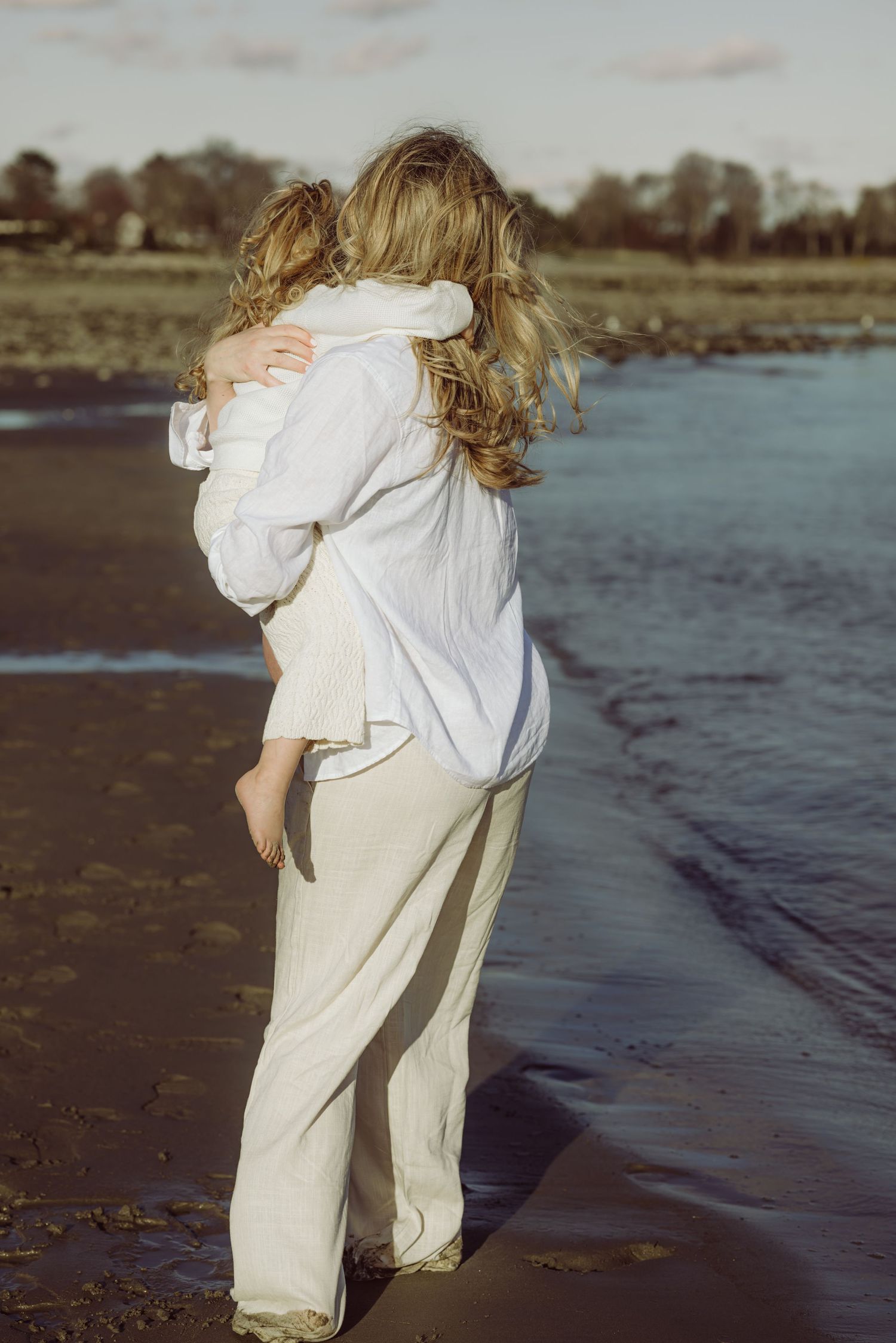 Heartwarming moment showing pregnant mother & daughter in white linen embracing on a Connecticut beach in Westport at sunset.