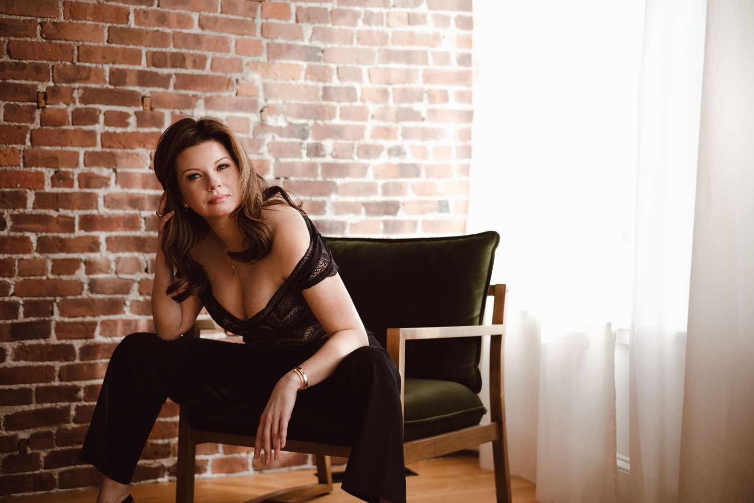 A woman in black lace top and black pants poses on a green suede chair against an exposed brick wall with natural window lighting.