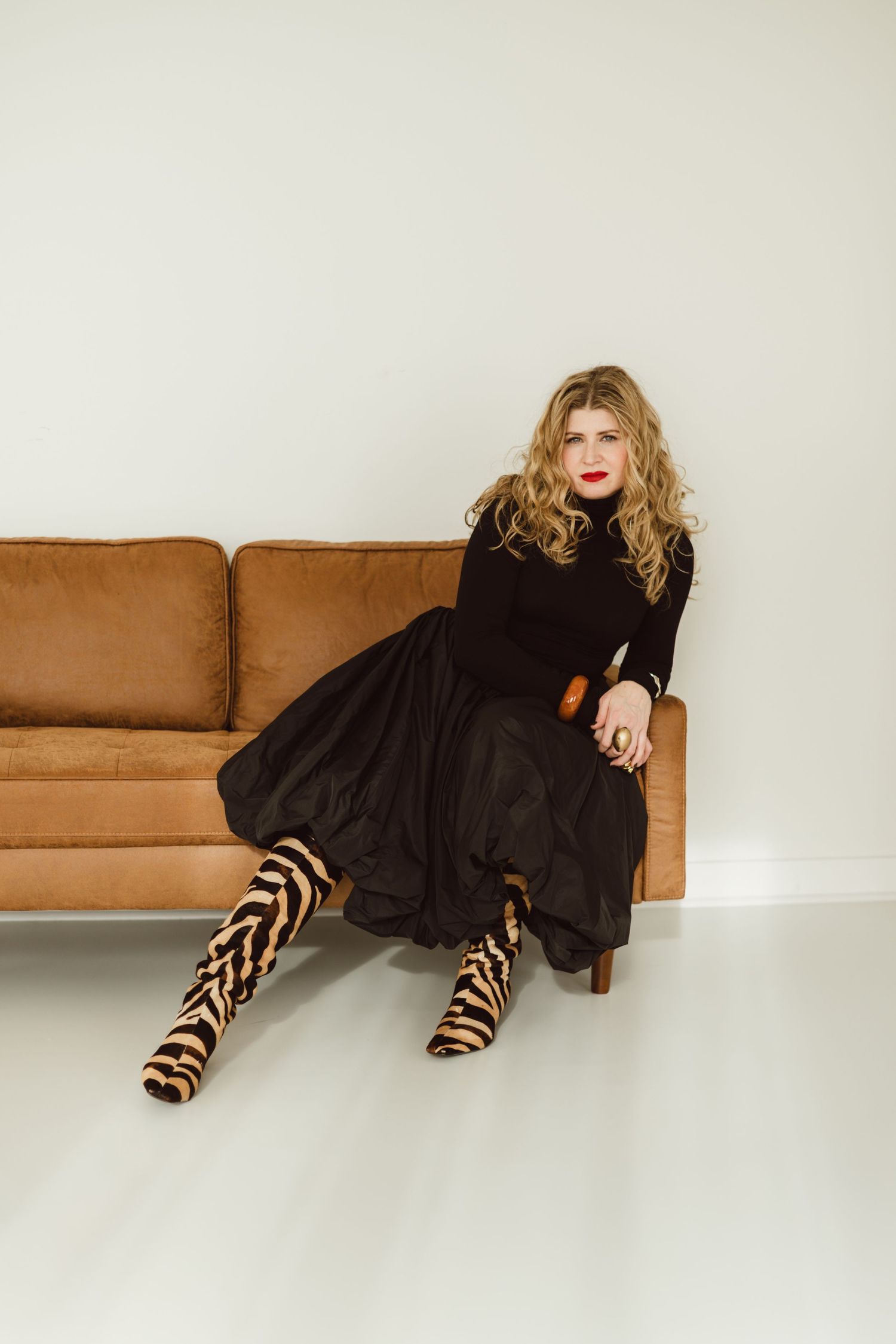 A women in a black dress reclines on a tan leather sofa against a white wall in a fashion editorial style pose with animal print high boots.