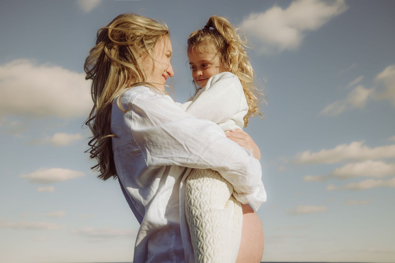 Two people in white clothing embrace tenderly against a cloudy sky backdrop on a bright sunny day.