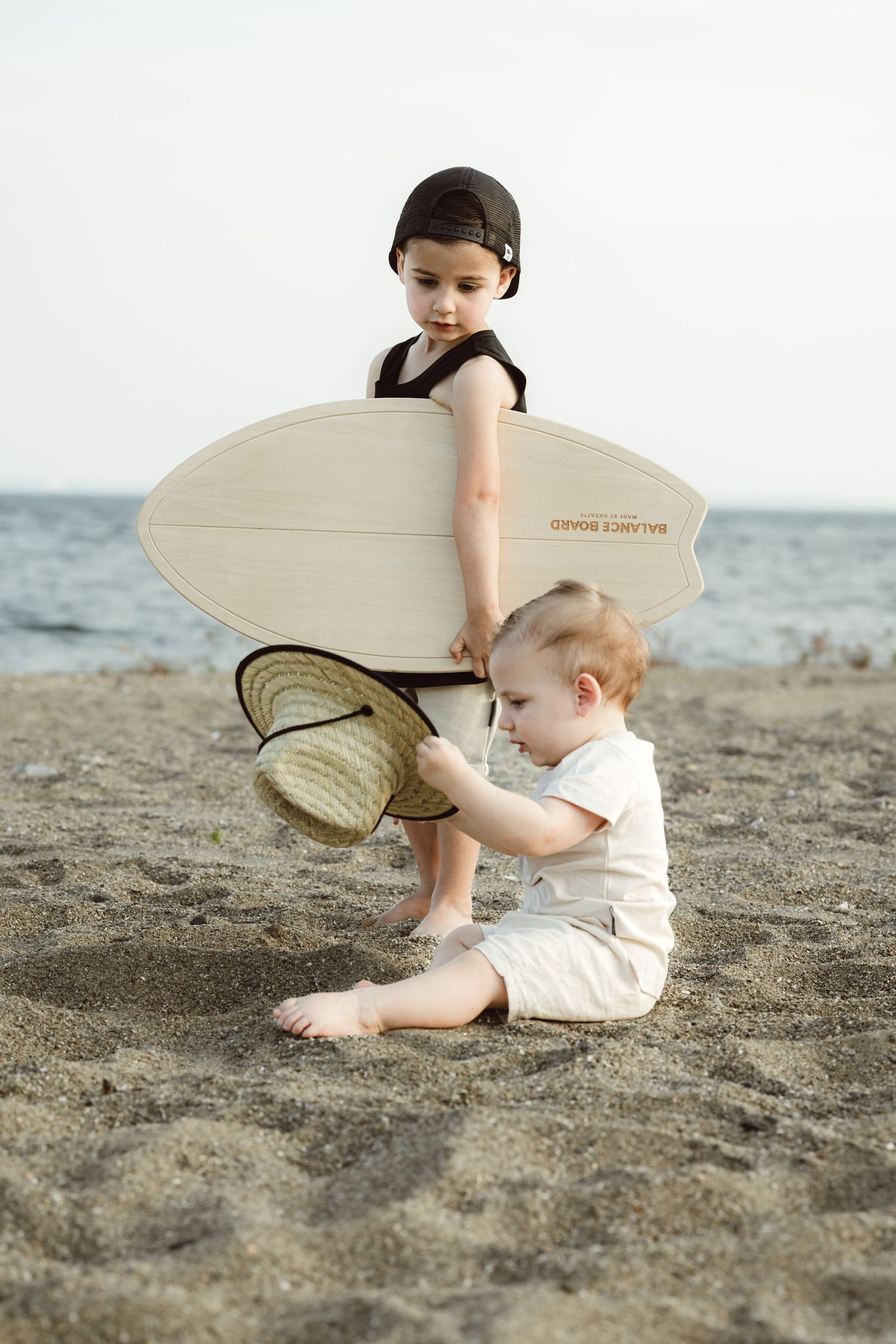 A young child sits on a sandy beach with a beach hat while toddler brother stand above holding a surfboard, creating a serene coastal scene.