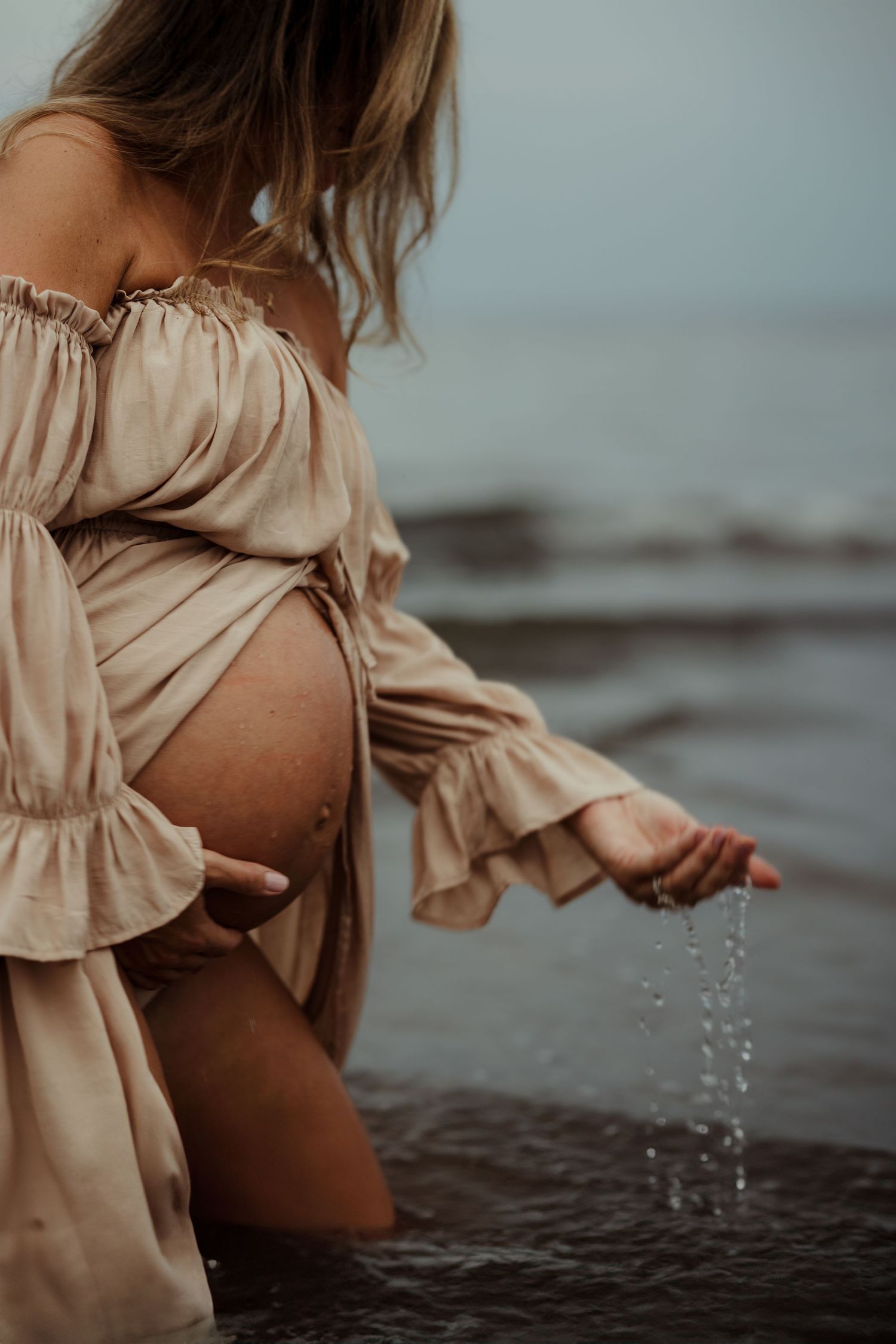 A pregnant woman in a beige dress plays with water droplets at the beach during sunset, creating a dreamy atmosphere.