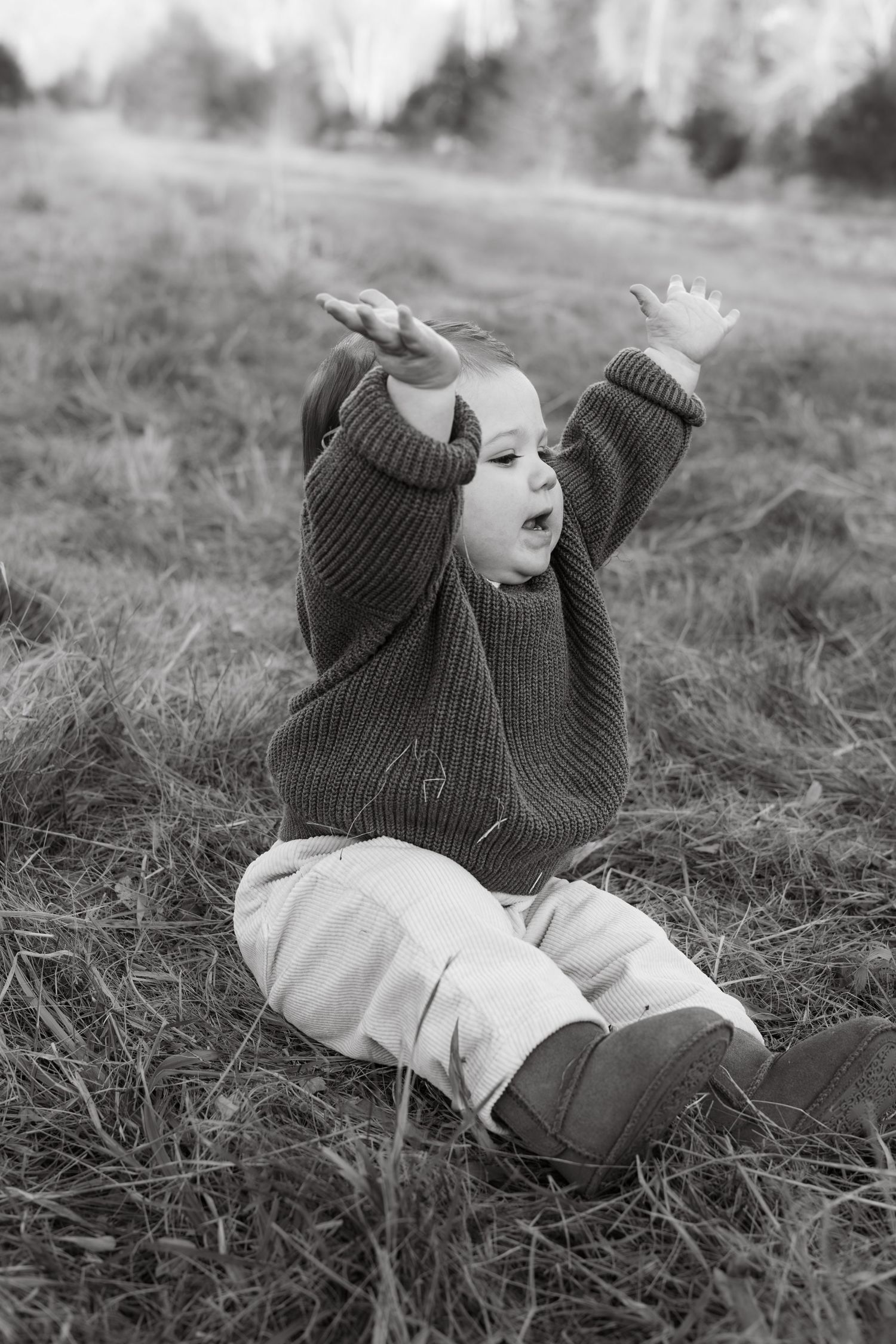 Black and white photo of a one year old girl with her hands raised to the sky asking for uppy from her mom during milestone photoshoot.
