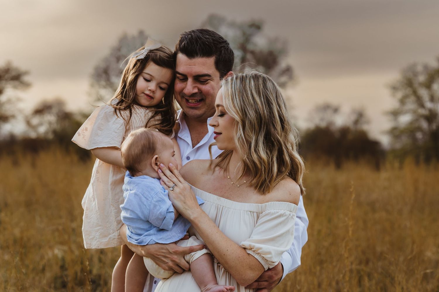 Family wearing white outfits embraces while standing in a golden field during sunset.