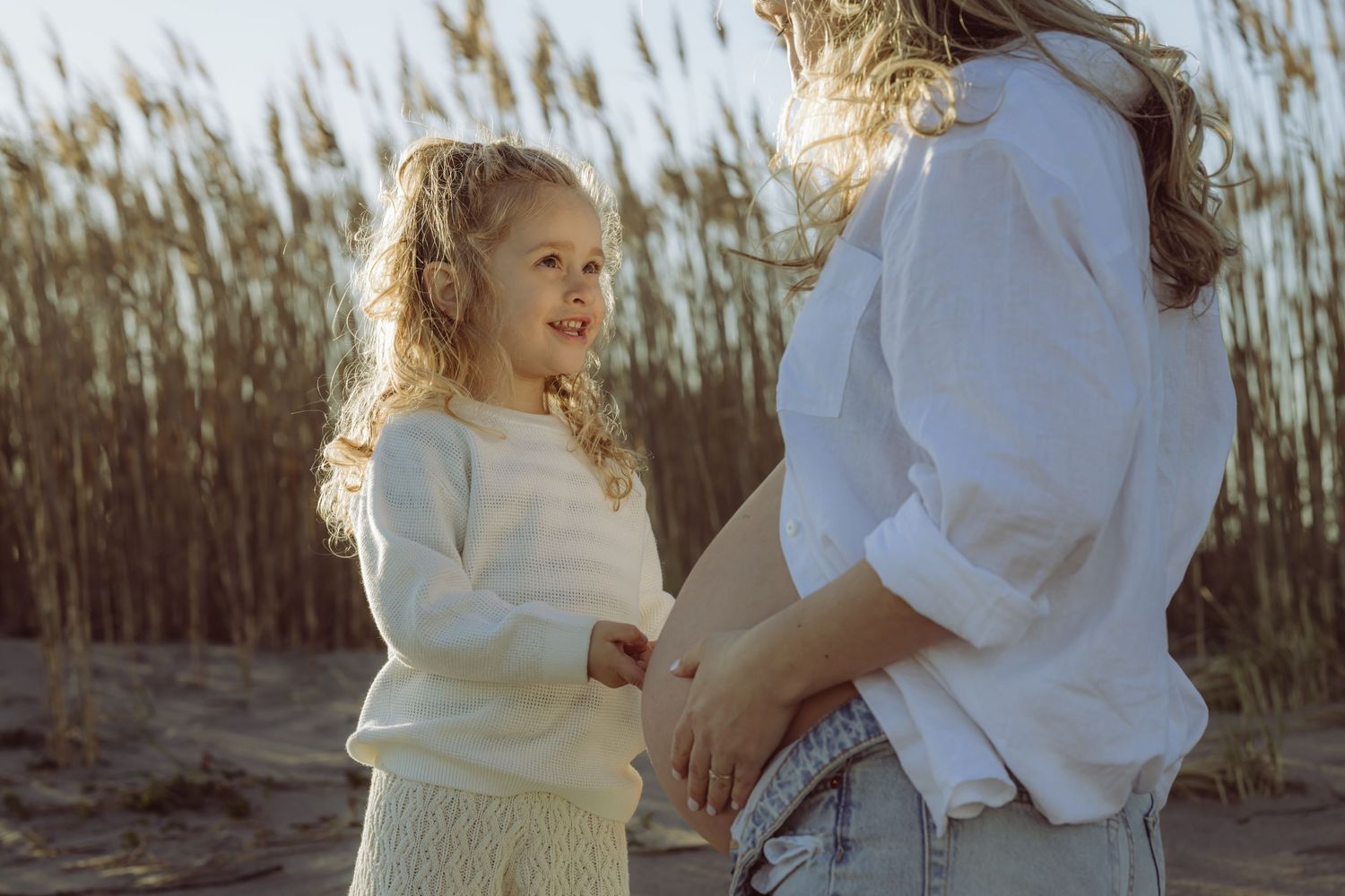 A heartwarming moment between parent and child standing together against tall reeds during golden hour. Daughter touching pregnant belly.