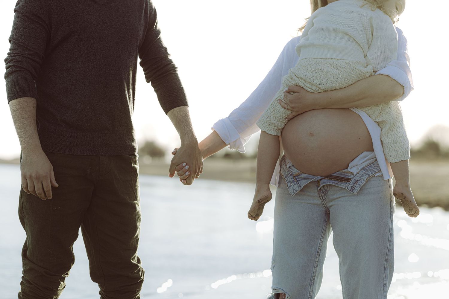 Pregnant woman holding daughter holding hands with husband walking on a sandy beach at sunset with golden sunlight in the background.