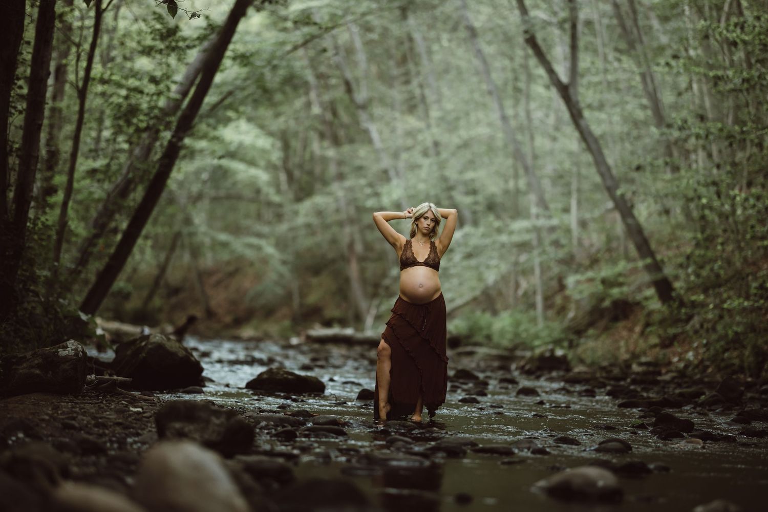 A pregnant woman stands in a tranquil forest stream that flows through mossy rocks for maternity photoshoot