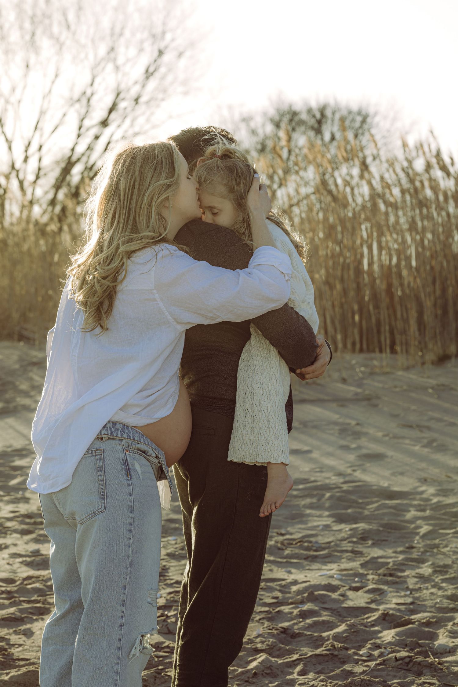 A pregnant mother embraces and kiss daughter on a sandy beach at sunset with bare trees in the background. Husband is holding her daughter.
