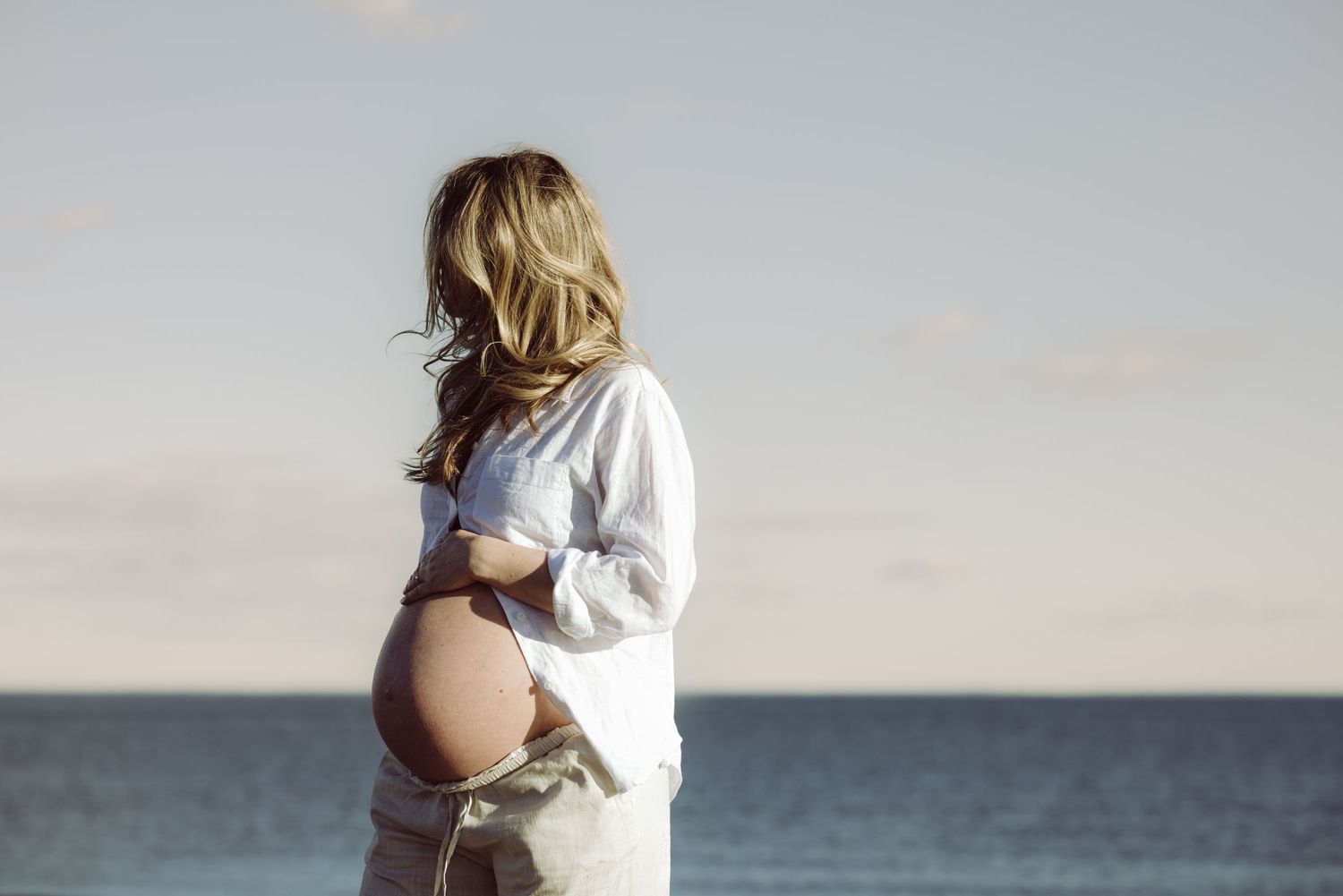 A pregnant woman in a white linen set stares into the ocean distance as she holds her pregnant belly.