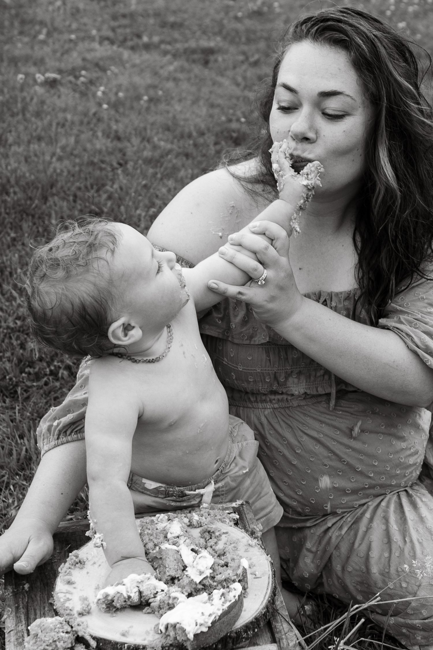 A mother and baby share cake together outdoors in a candid black and white photograph.