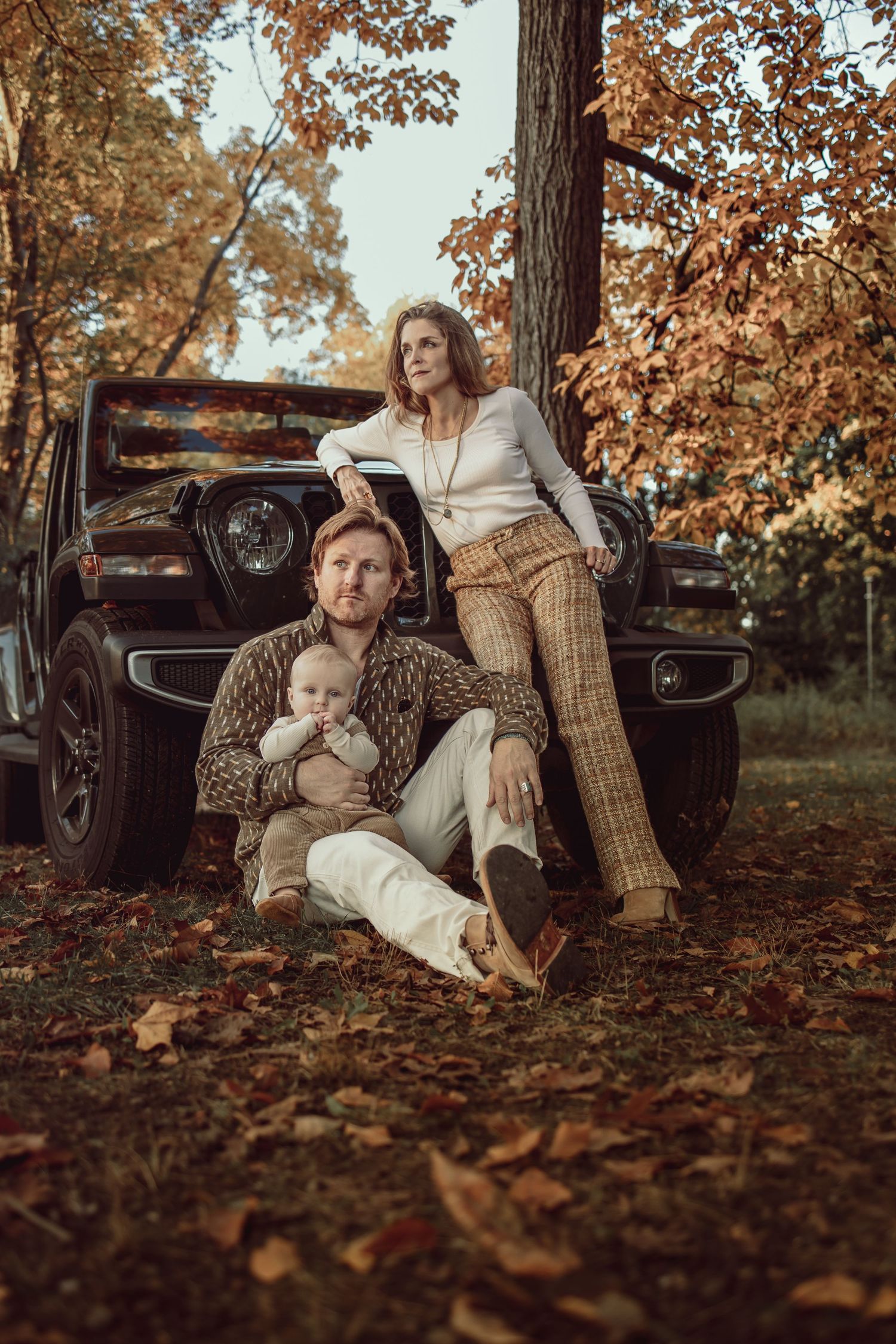 Cozy autumn family portrait with vintage car surrounded by fallen leaves and warm brown tones in a forest setting.