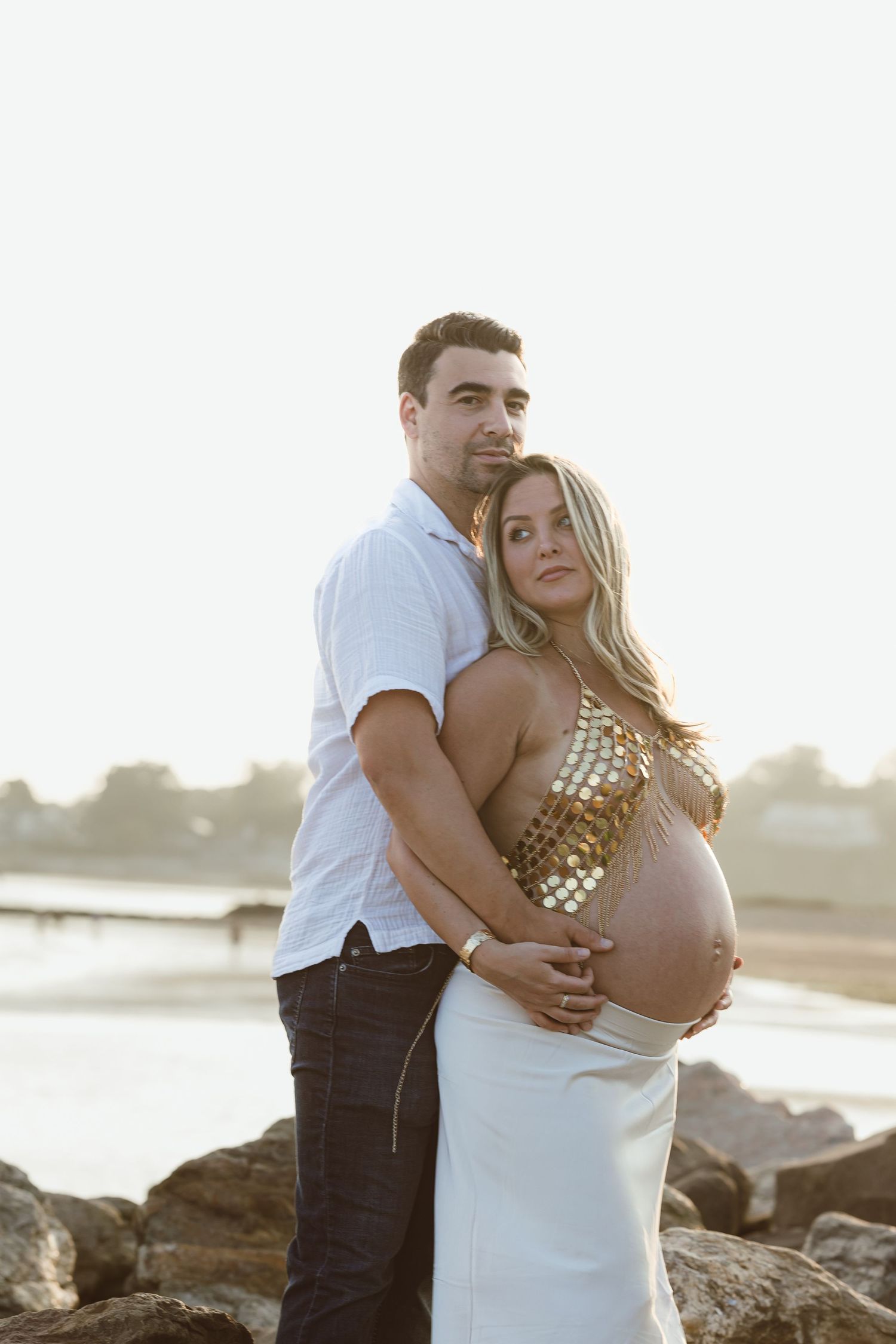 A couple embraces during a romantic maternity sunset photoshoot on a rocky beach.