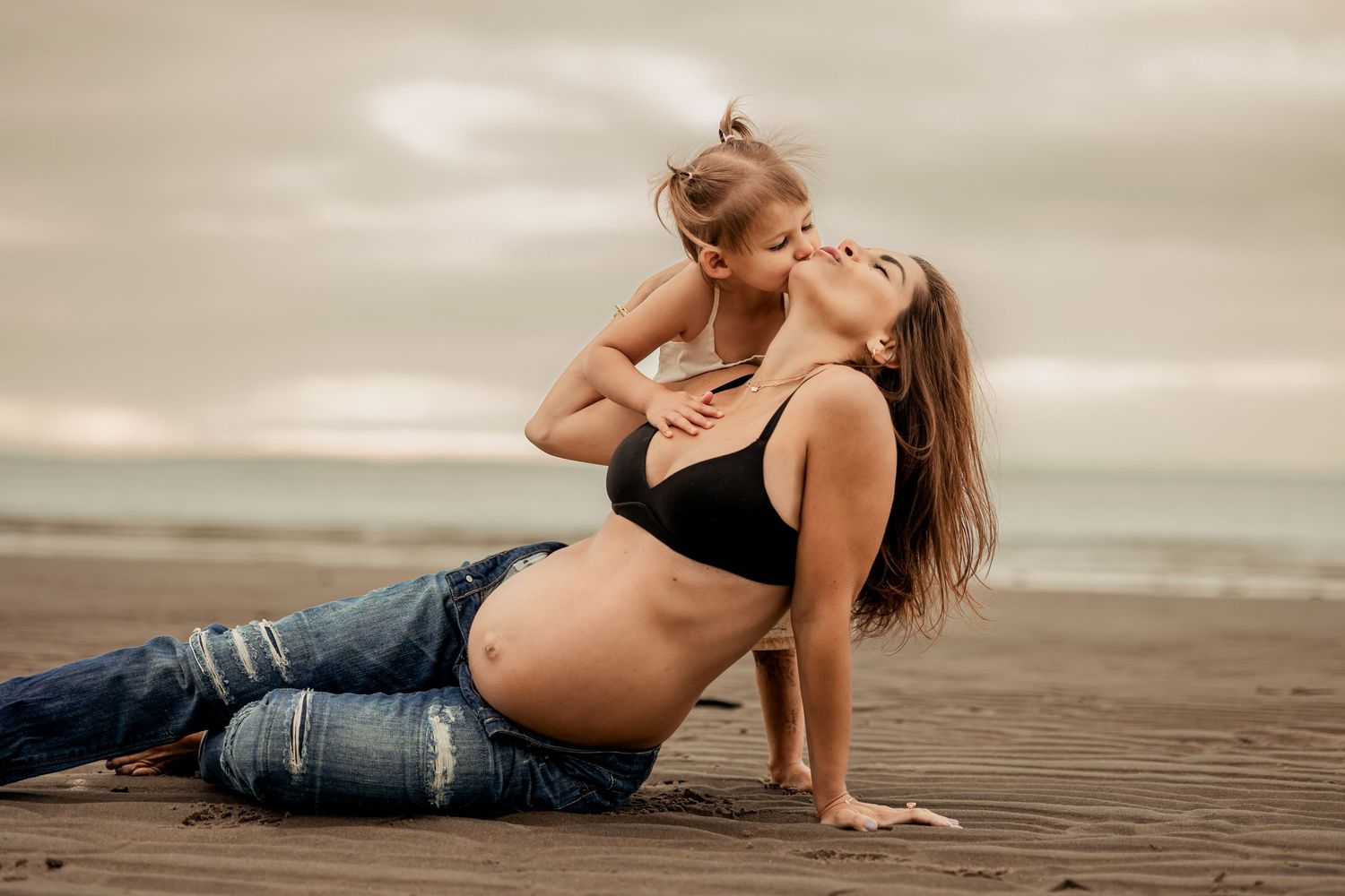 Woman in black bikini top and jeans poses gracefully for a maternity photoshoot on the beach at sunset with her daughter kissing her cheek.
