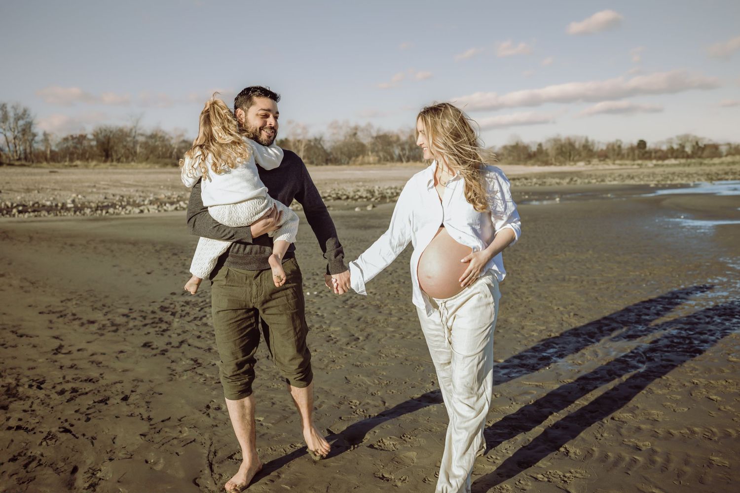 A playful couple enjoys a sweet and romantic moment during a maternity photoshoot on a sandy beach at sunset with their toddler daughter.