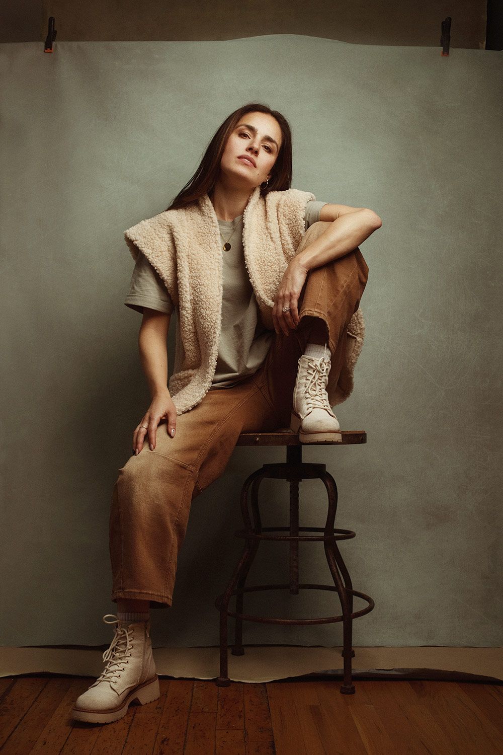 Artistic fashion portrait of a woman photographer in beige and brown tones posing with a wooden chair against textured wall.