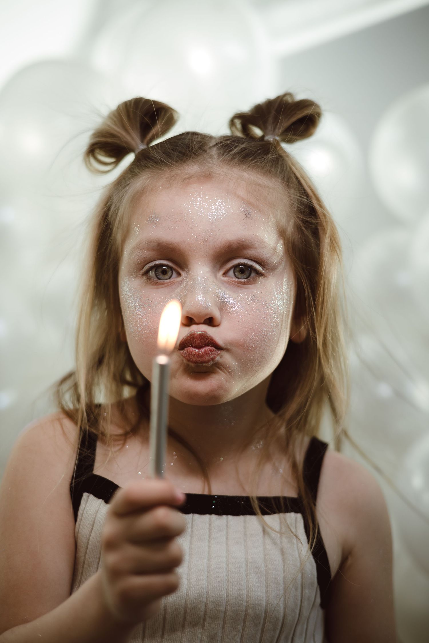 A child with pigtails holds a lit birthday candle while making a puckered expression against a soft blurred background for photoshoot.