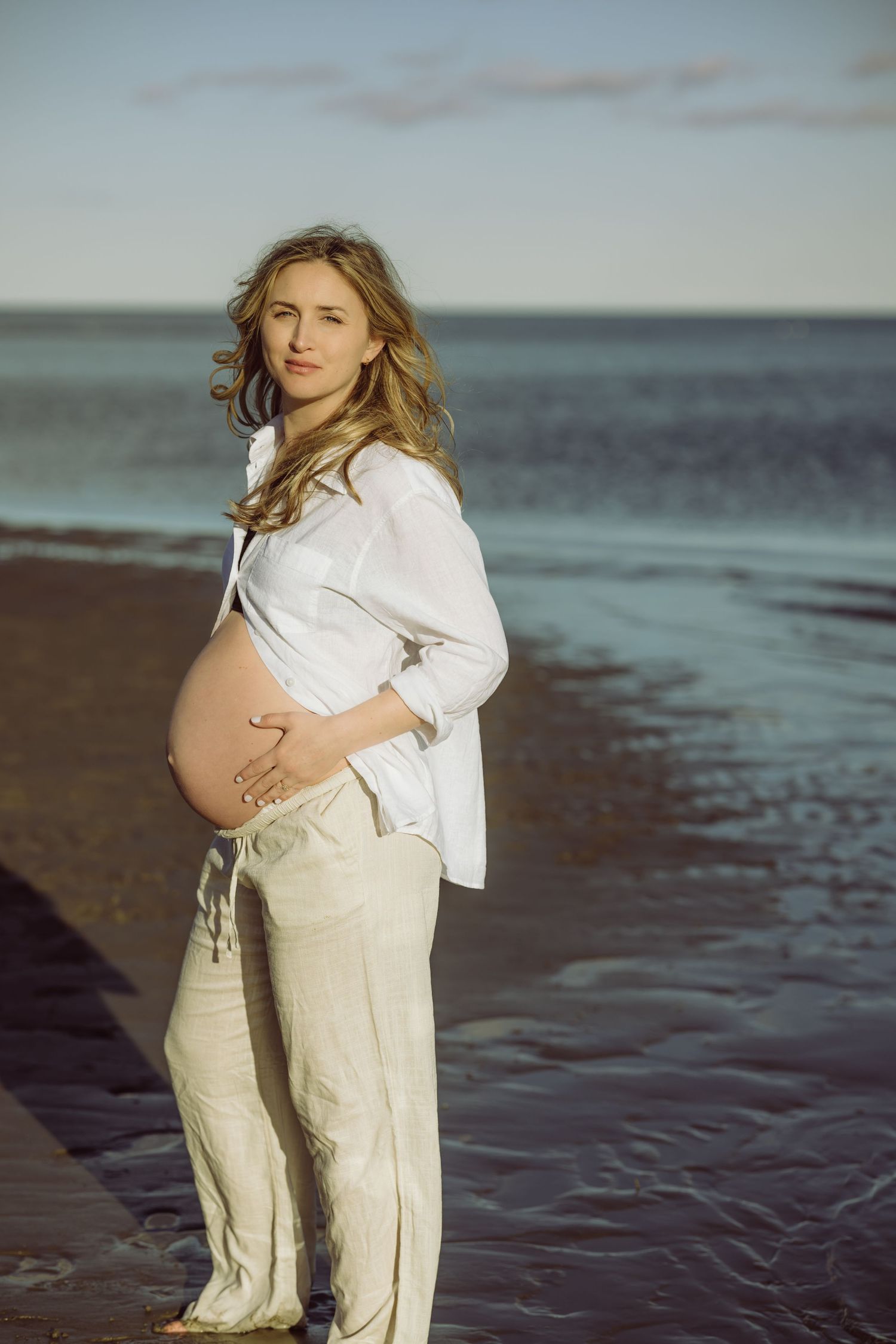 Pregnant woman in white shirt and beige pants stands on beach at sunset with ocean waves in background for maternity session.
