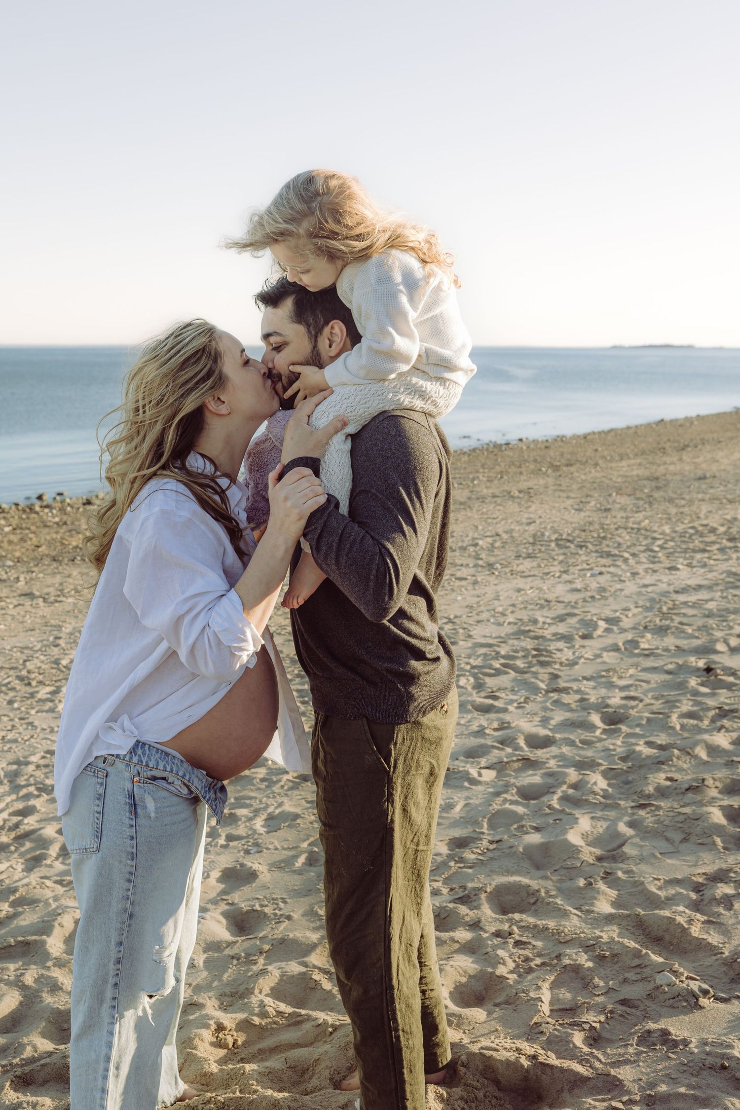 Pregnant woman, husband and toddler girl enjoying a tender moment together on sandy westport, CT beach at sunset with ocean in background.