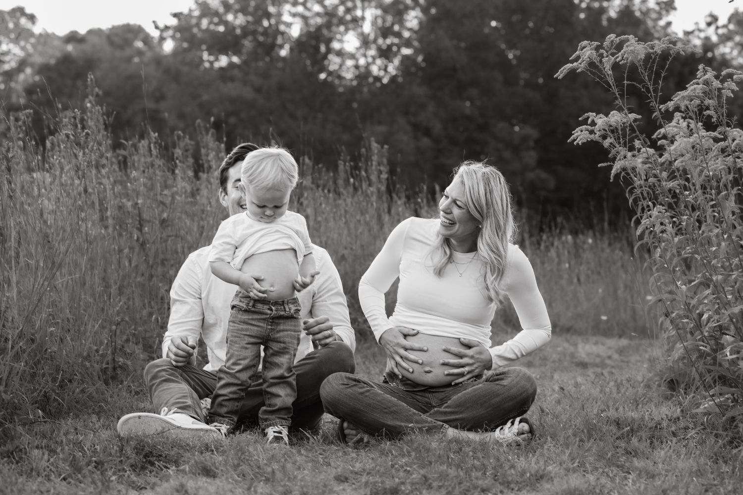 A couple sits in the grass. The mother is pregnant holding her belly and laughs as her toddler son stands and does the same.
