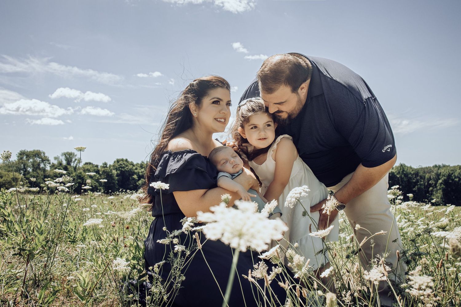 A couple shares a tender moment in a blooming meadow while surrounded by white wildflowers and summer clouds and their two children.