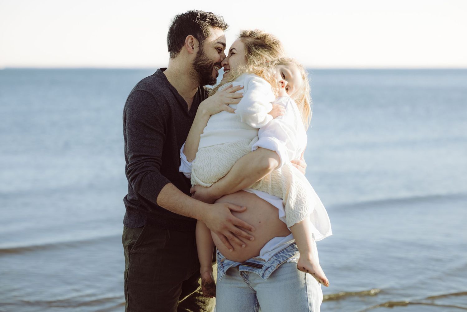 Father gently touches wifes pregnant belly while mother holds toddler daughter on a Westport beach during maternity photoshoot.