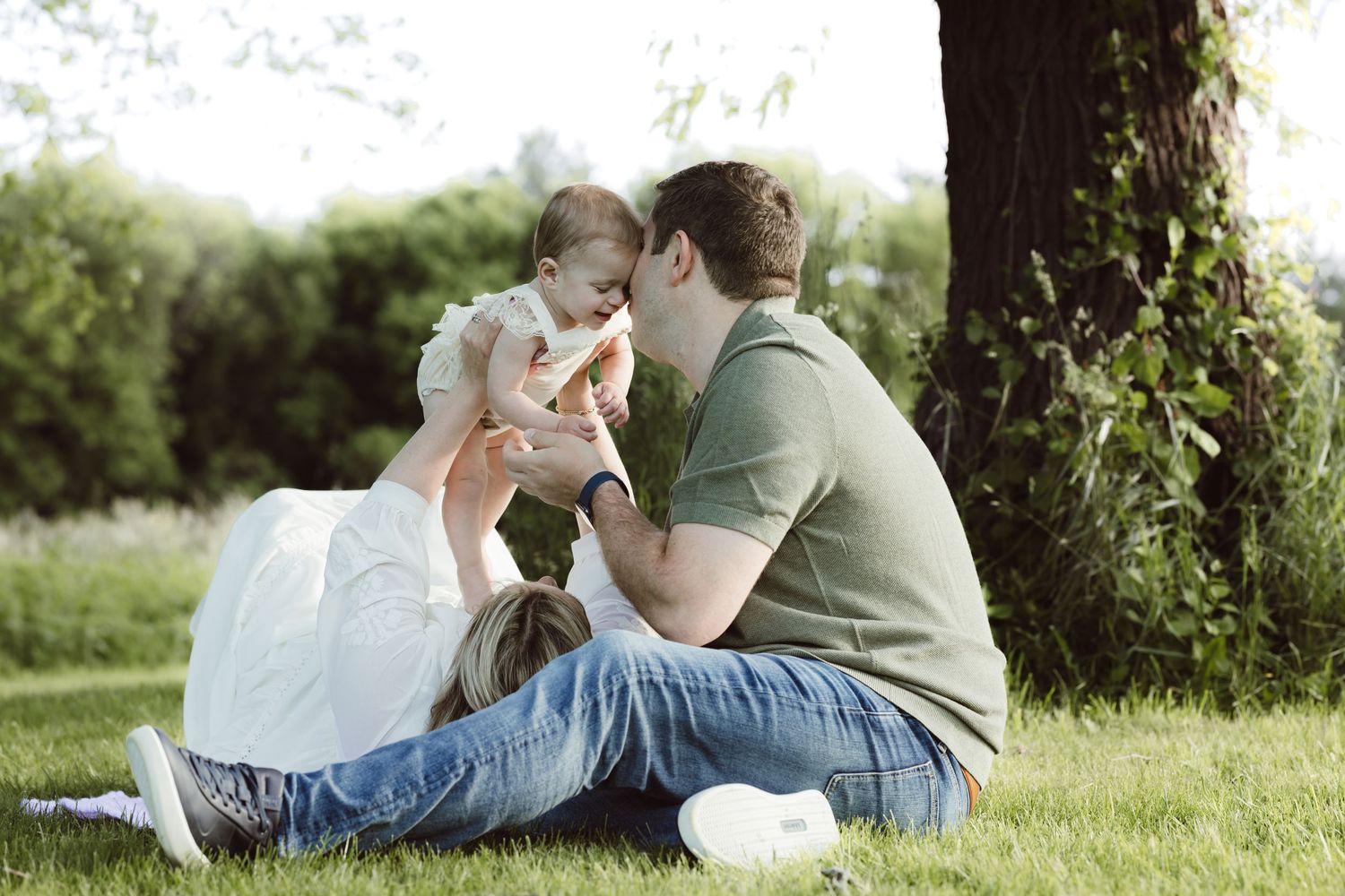 Couple relaxing together on grass under a tree during a sunny summer afternoon holding their child above their heads kissing her forehead.