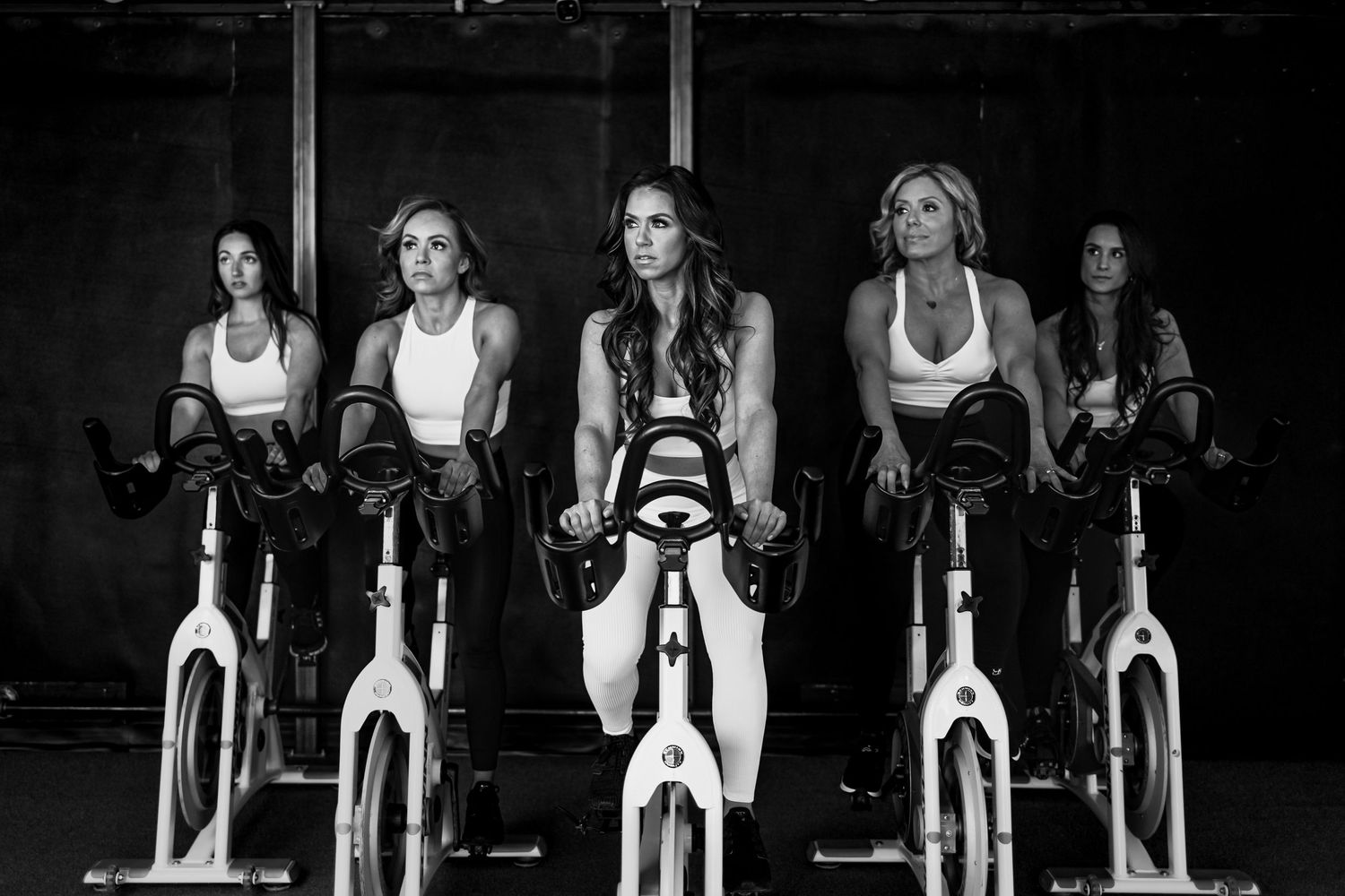A group of women trainers exercising on stationary spin bikes in a dark fitness studio during a cycling class for fitness photoshoot.