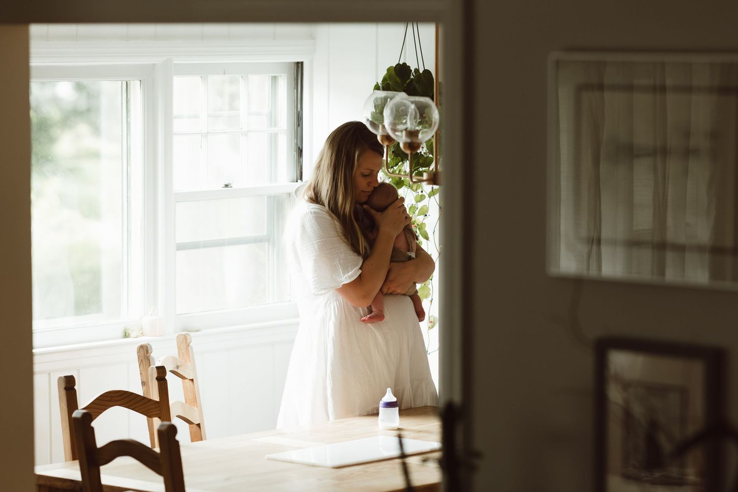 A mother in a white dress stands by a window in kitchen in soft natural light, creating a dreamy silhouette holding newborn baby girl.