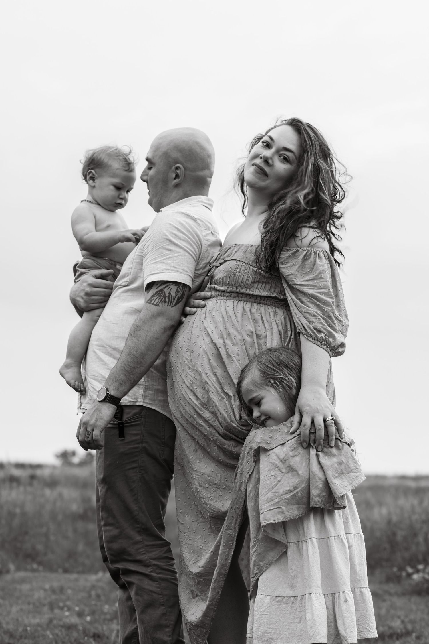 Black and white photo showing a loving family of 4 embrace in a field with candid emotional moments during a maternity session.