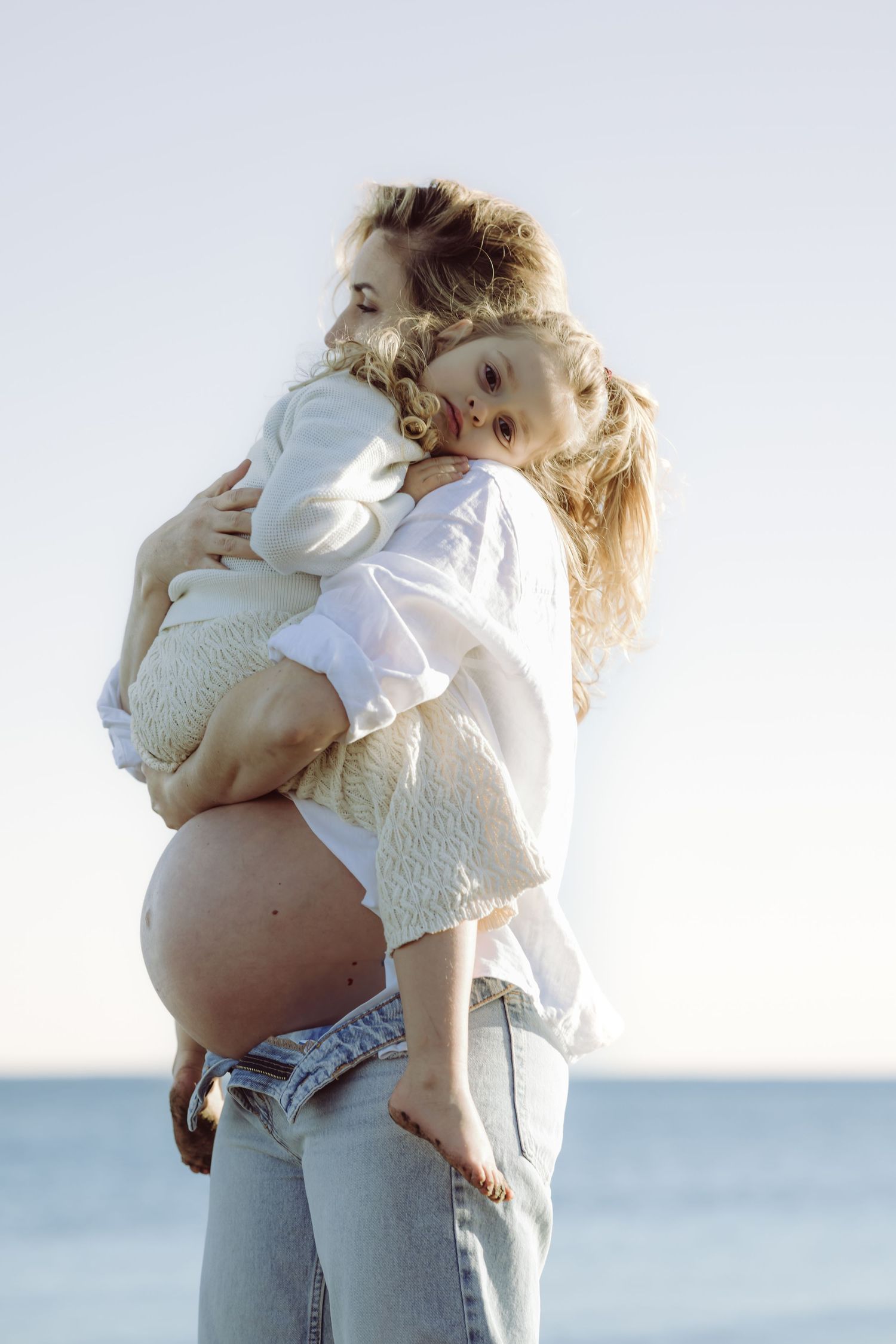 Pregnant Mother & Daughter in white linen embrace tenderly by the water at sunset on Sherwood island in westport.